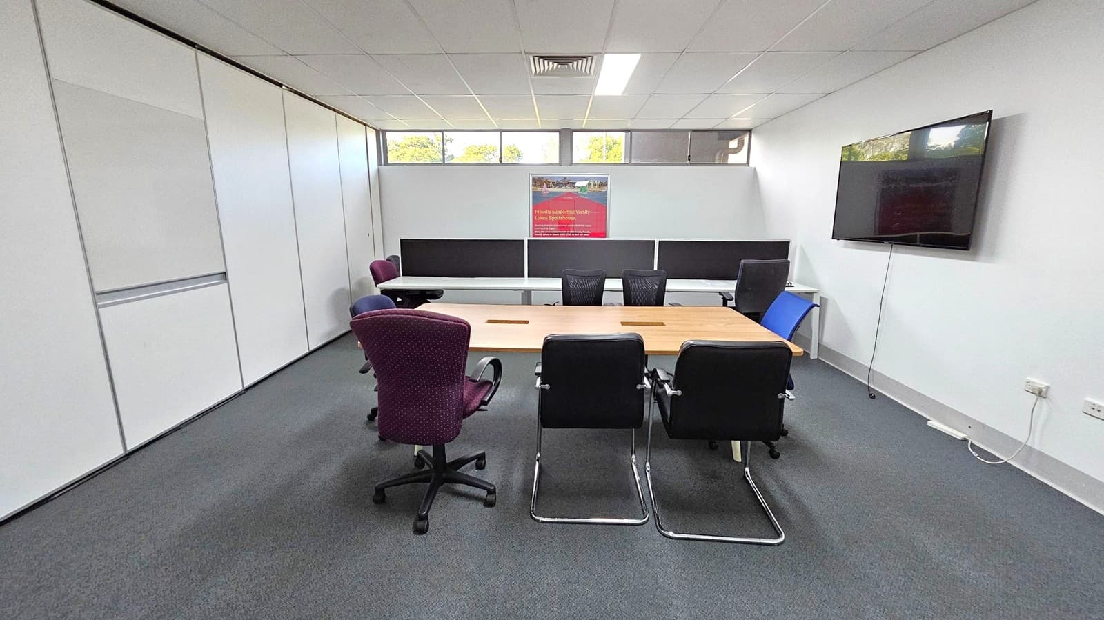 A conference room with a rectangular wooden table in the centre, surrounded by various chairs, including black, blue, and maroon ones. On the right wall, a large flat-screen TV is mounted. The back wall features a poster and windows above, allowing natural light. The left wall is lined with white panels. The floor is carpeted in grey, and the ceiling is fitted with white tiles and fluorescent lights.