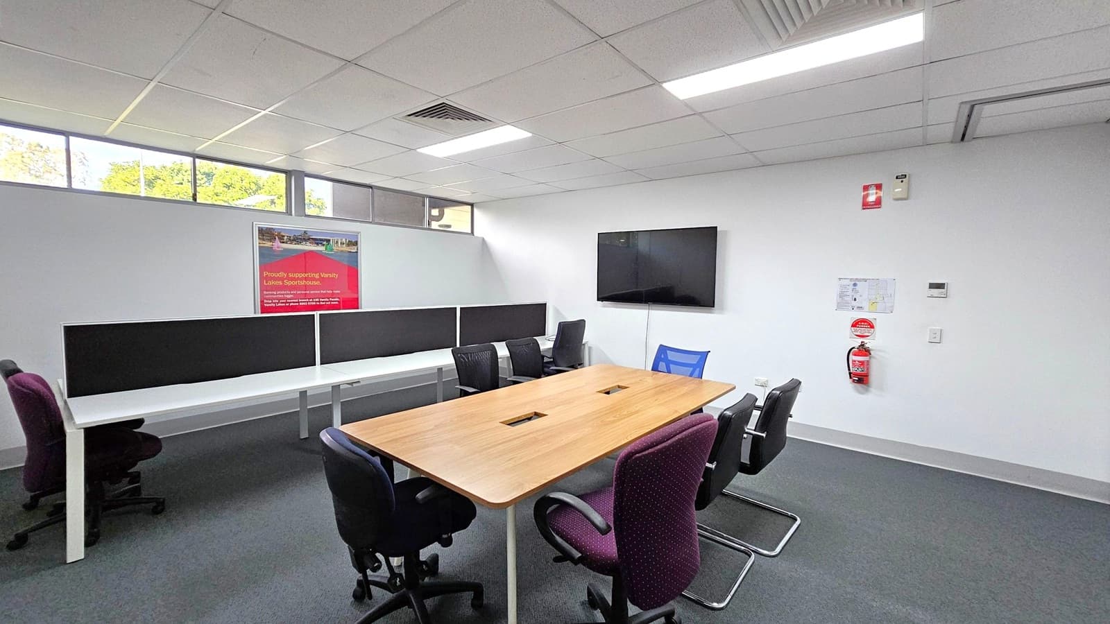 A modern office space with a rectangular wooden table in the centre, surrounded by various office chairs. On the left, there are white desks with black dividers and additional chairs. A red poster is mounted on the wall above the desks. On the right, a flat-screen TV is mounted on the wall, with a fire extinguisher and a small map below it. The ceiling is fitted with fluorescent lights and air vents, and the floor is carpeted in grey. High windows allow natural light in.