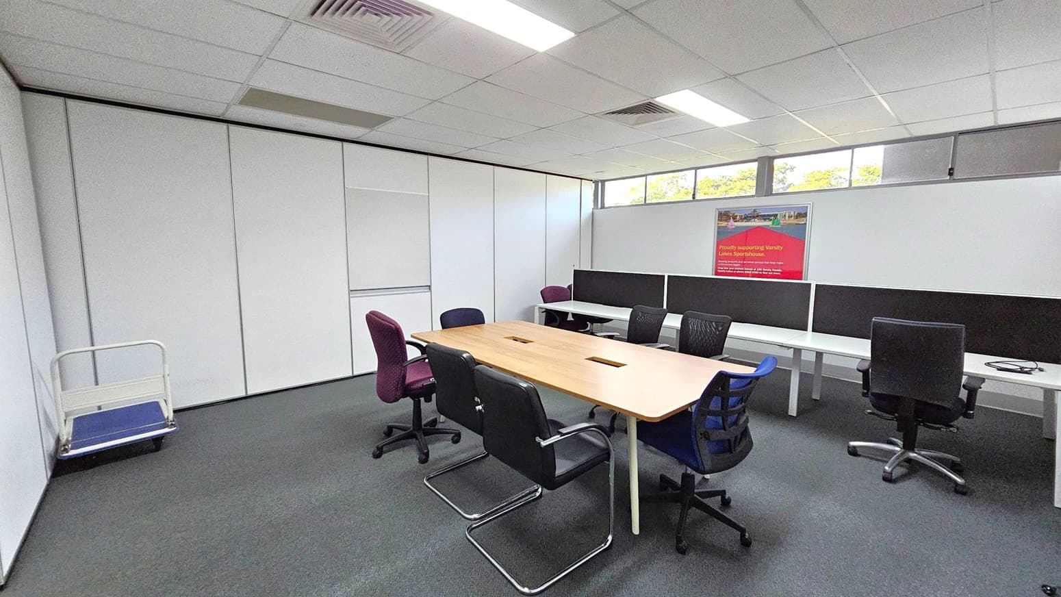 A conference room with a long wooden table in the centre surrounded by various office chairs, including black, purple, and blue ones. On the right, there are white desks with black dividers and additional chairs. A poster is mounted on the wall above the desks. The left side has a small trolley against a white partition wall. The ceiling is fitted with fluorescent lights and air vents, and the floor is carpeted in grey. High windows near the ceiling let in natural light.