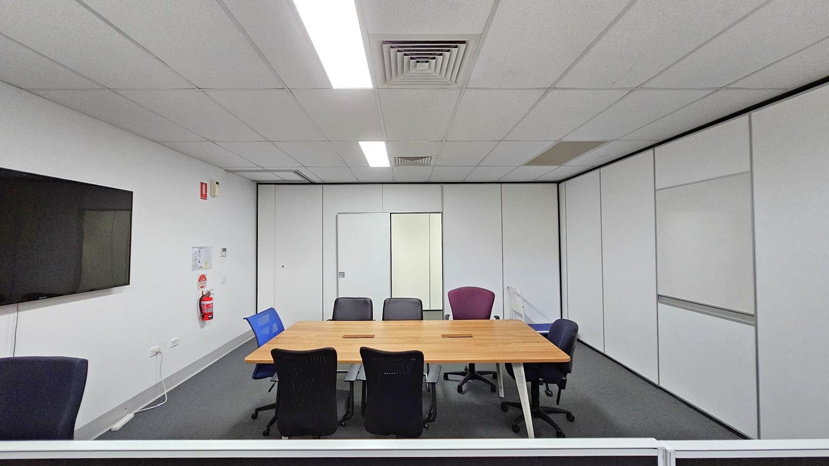 A conference room with a rectangular wooden table in the centre, surrounded by various office chairs. On the left wall, a large flat-screen TV is mounted, with a fire extinguisher and some notices below it. The walls are white with a door in the centre. The ceiling has fluorescent lights and air vents, and the floor is carpeted in grey.