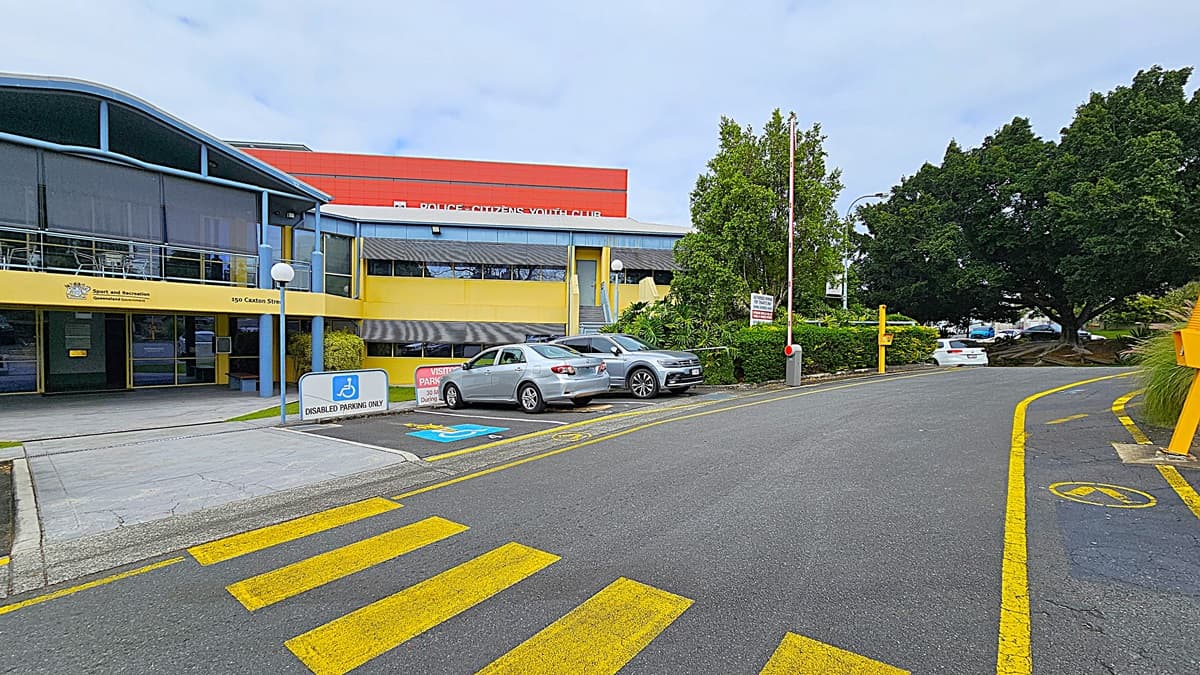 A two-storey building with a blue and yellow facade is in the centre, labelled "Sport and Recreation." The foreground has a paved area with yellow pedestrian crossing lines and a disabled parking sign. On the left, there is a concrete walkway leading to the building entrance. On the right, there are parked cars, including a silver sedan and a grey SUV, with a barrier arm and greenery. The background includes a red building with partial signage and dense green trees.