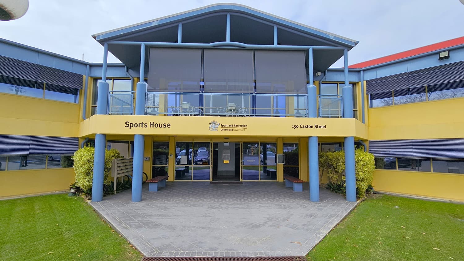 A two-storey building with a blue and yellow facade is in the centre, labelled "Sports House" with the address "150 Caxton Street." The foreground has a tiled walkway leading to the entrance, flanked by blue pillars. On the left, there are green bushes and a small bench. On the right, another bench and more greenery. The background includes large windows with blinds on both floors. The floor is tiled at the entrance and surrounded by a well-maintained grass lawn.