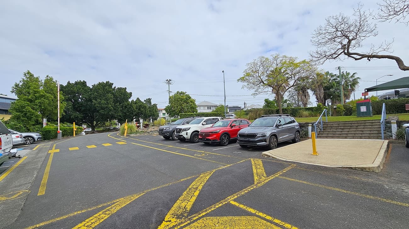 On the left, a paved driveway with yellow pedestrian crossing lines curves past a barrier arm and green trees. In the middle, a row of parked cars includes a white SUV, a red car, and a grey SUV. On the right, a concrete ramp and steps with blue railings lead up to a grassy area with a bench and a sign. The background features more greenery, including palm trees and a few residential buildings. The floor is asphalt with marked yellow lines.