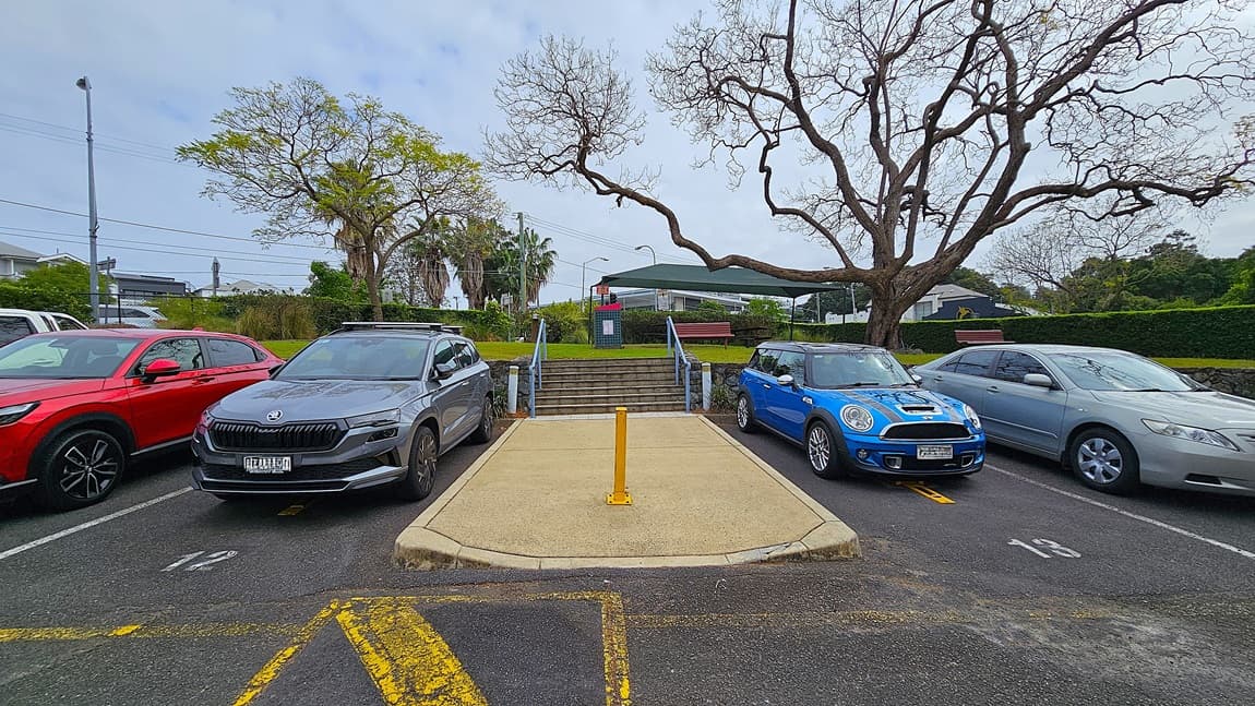 On the left, a red SUV and a grey SUV are parked in marked spaces. In the middle, a concrete ramp with a yellow bollard leads to a set of stairs with blue railings. On the right, a blue Mini Cooper and a silver sedan are parked. The background features a green lawn, a bench, and a covered structure with a red sign. Leafless trees with sprawling branches frame the scene. The floor is asphalt with yellow parking lines and a concrete ramp.
