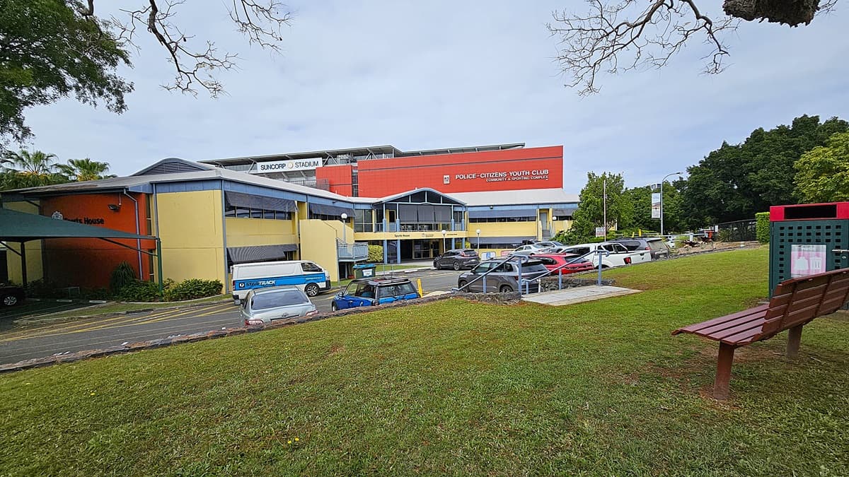 On the left, a yellow and orange building with a sign reading "Sports House" and a blue van parked nearby. In the middle, a two-storey building with a blue and yellow facade labelled "Police-Citizens Youth Club" and "Suncorp Stadium" in the background. On the right, a grassy area with a wooden bench and a green bin. Several cars, including a blue hatchback and a white SUV, are parked in front. The floor is asphalt with marked parking spaces and a concrete walkway.
