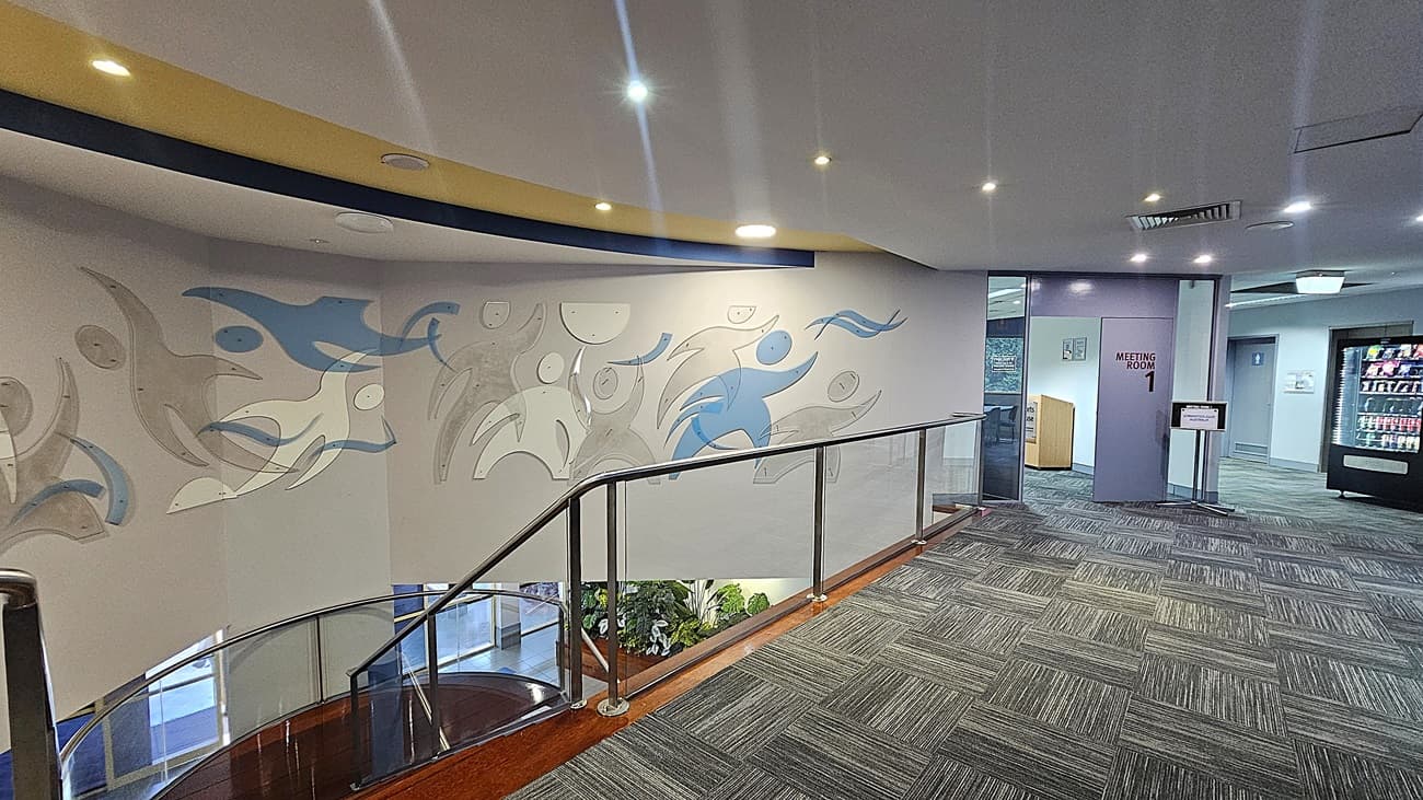 On the left, abstract figures in grey and blue on a white wall. In the middle, a staircase with wooden steps and glass railings leading down to a lower level with plants. On the right, a continuation of the abstract figures on the wall. The floor is covered with grey patterned carpet tiles. The ceiling is white with recessed lighting and a yellow and blue curved design element.