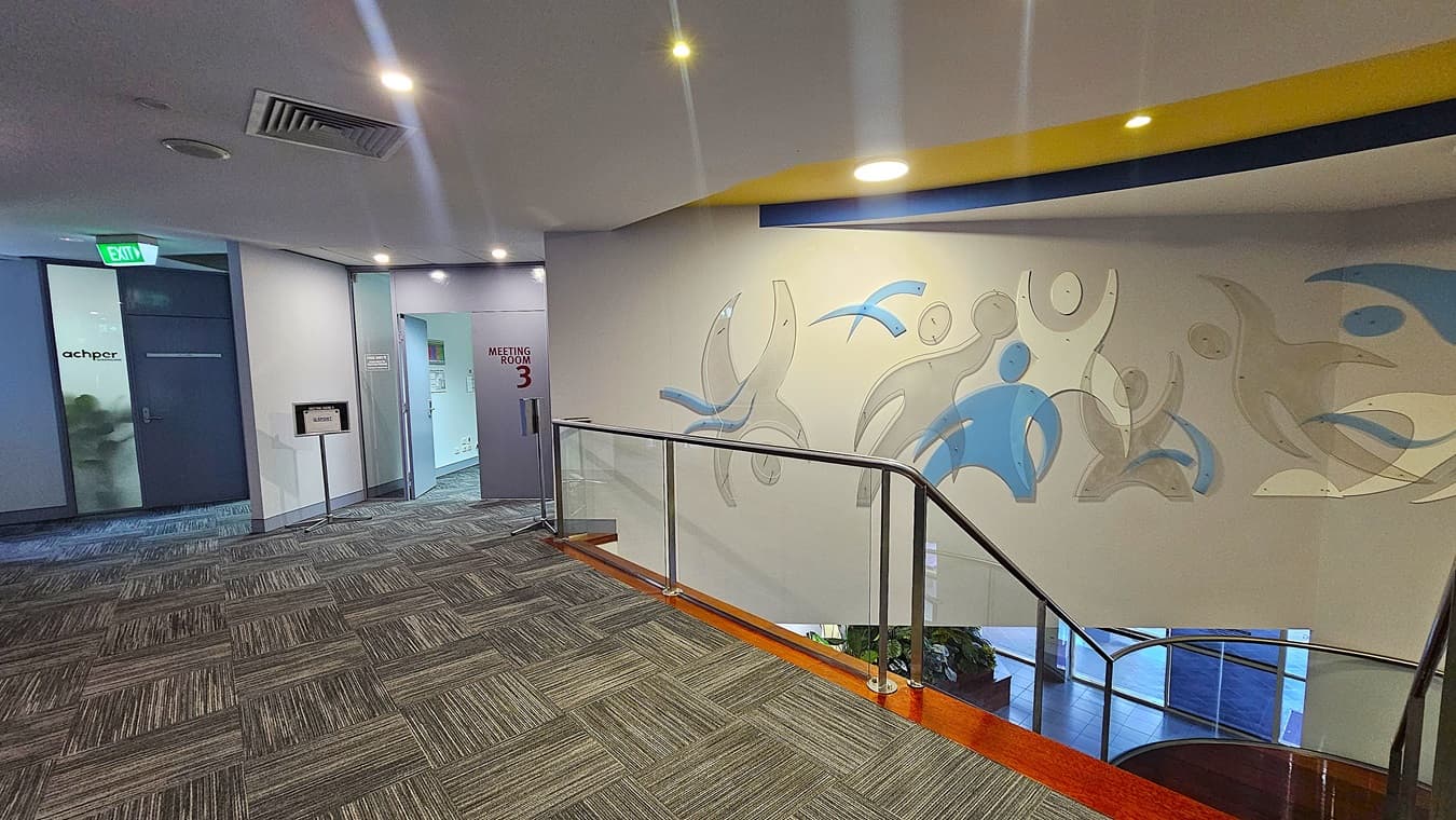 On the left, a frosted glass door with the "achper" logo and an emergency exit sign above it. In the middle, an open door labelled "Meeting Room 3" with a sign next to it. On the right, a white wall with abstract blue and grey figures above a staircase with glass railings and wooden steps. The floor is covered with grey patterned carpet tiles. The ceiling is white with recessed lighting and a yellow and blue curved design element.