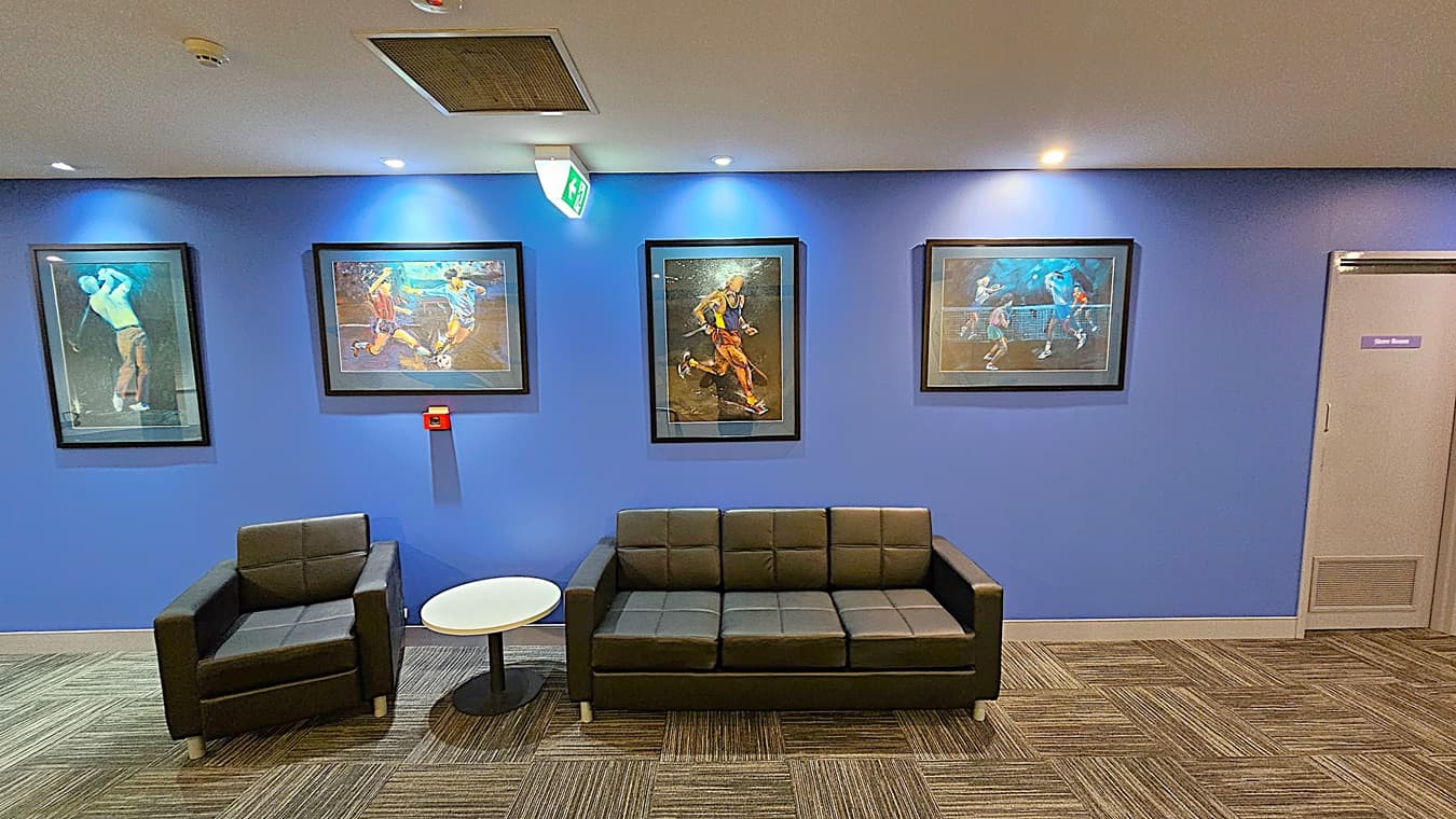 On the left, a black armchair next to a small round white table. In the middle, a black three-seater sofa beneath three framed sports paintings on a blue wall, with an emergency exit sign above. On the right, a grey door labelled "Store Room" and another framed sports painting. The floor is covered with grey patterned carpet tiles. The ceiling is white with recessed lighting and a large air vent.