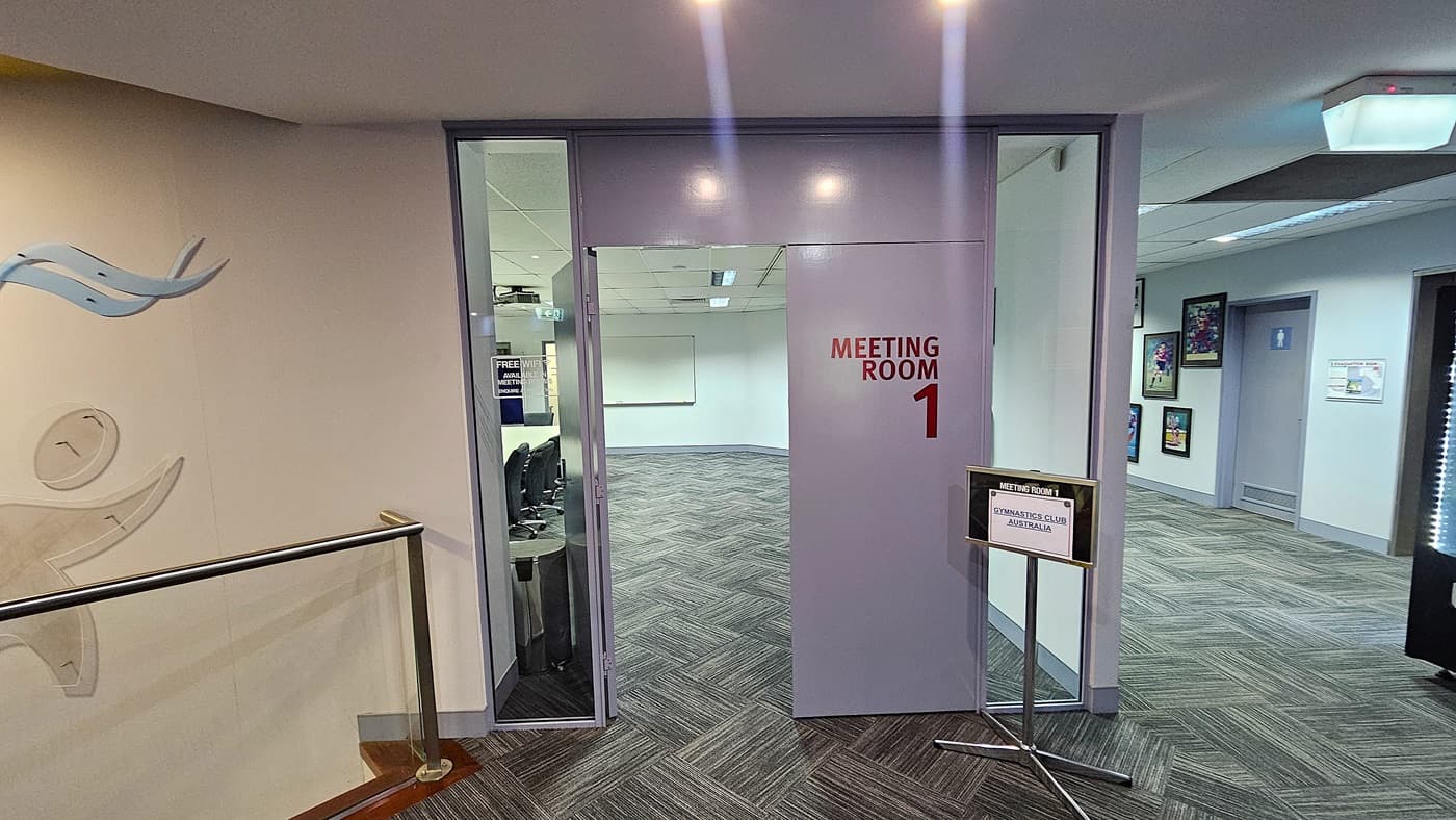On the left, a wall with abstract blue and grey figures above a staircase with glass railings and wooden steps. In the middle, an open door labelled "Meeting Room 1" leading to a room with grey patterned carpet tiles and a whiteboard on the far wall. On the right, a hallway with framed pictures on the walls and a sign reading "Gymnastics Club Australia." The floor is covered with grey patterned carpet tiles. The ceiling is white with recessed lighting and air vents.