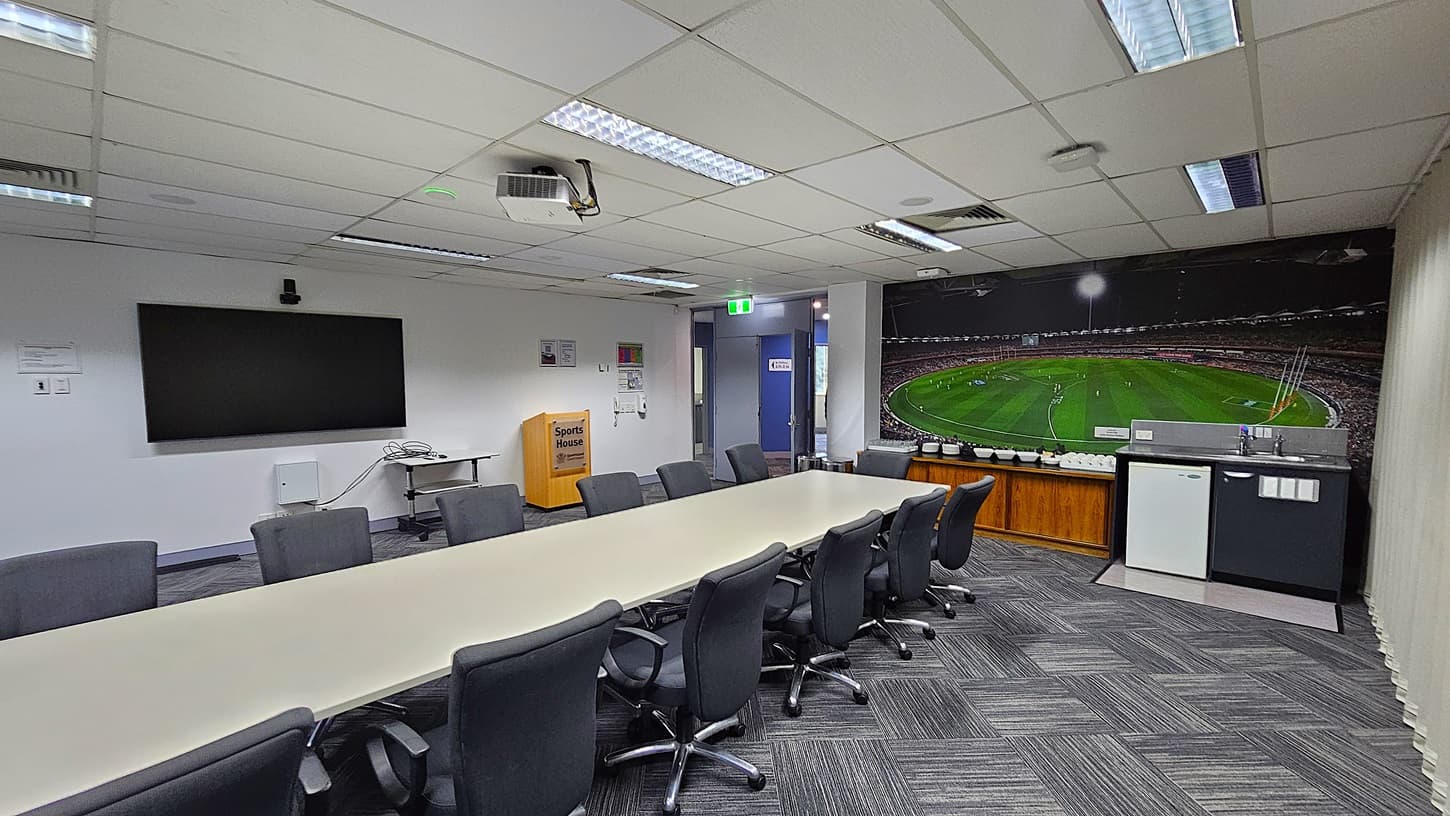 On the left, a large wall-mounted TV above a small table with cables. In the middle, a long rectangular table surrounded by black office chairs, with a ceiling-mounted projector above. On the right, a wall-sized mural of a sports stadium above a counter with a mini-fridge and a sink. The floor is covered with grey patterned carpet tiles. The ceiling is white with fluorescent lighting, air vents, and acoustic tiles.
