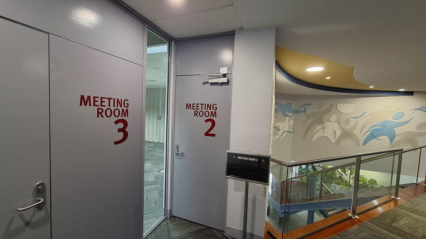 On the left, a closed door labelled "Meeting Room 3" in red text. In the middle, an open door labelled "Meeting Room 2" leading to a room with grey patterned carpet tiles. On the right, a staircase with glass railings and wooden steps, next to a wall with abstract blue and grey figures. The floor is covered with grey patterned carpet tiles. The ceiling is white with recessed lighting and a curved yellow and blue design element.