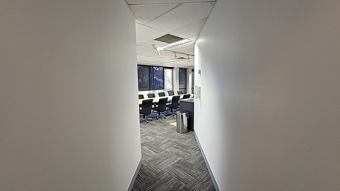 On the left, a plain white wall. On the right, another white wall with a small control panel and a metal bin near the entrance. In the middle, a long rectangular table surrounded by black office chairs, with large windows in the background. The floor is covered with grey patterned carpet tiles. The ceiling is white with recessed lighting, air vents, and acoustic tiles.