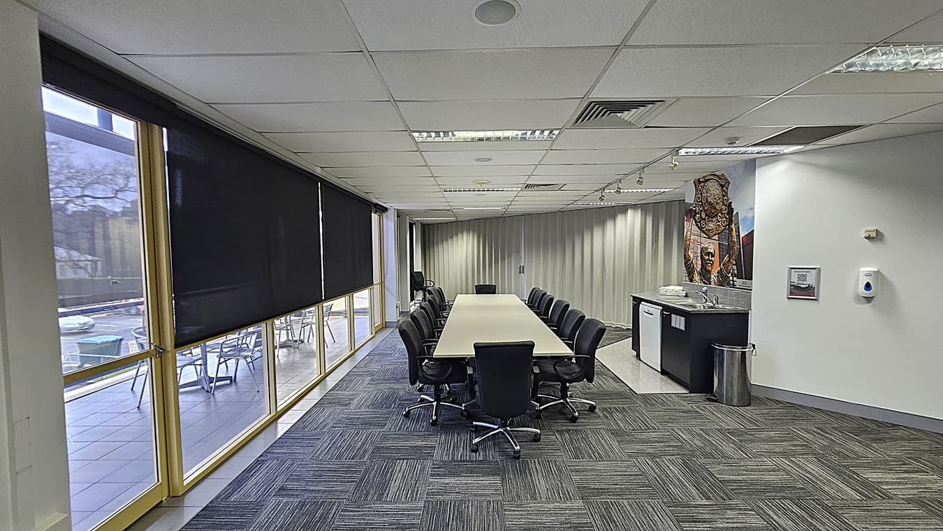 On the left, large windows with black roller blinds partially drawn, overlooking an outdoor seating area. In the middle, a long rectangular table surrounded by black office chairs. On the right, a counter with a sink, mini-fridge, and a mural of a historical figure above it, along with a metal bin and a wall-mounted hand sanitiser dispenser. The floor is covered with grey patterned carpet tiles. The ceiling is white with recessed lighting, air vents, and acoustic tiles.
