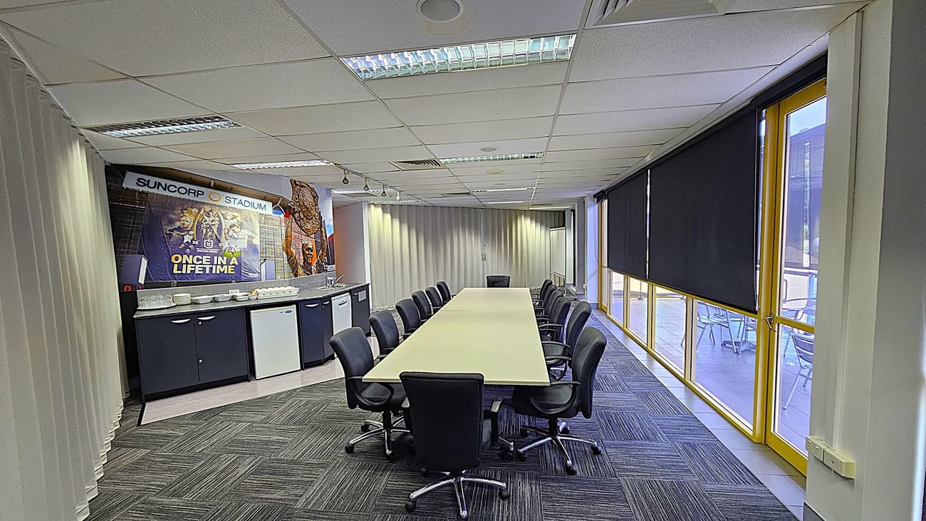 On the left, a counter with a sink, mini-fridge, and cabinets, with a mural of sports figures and the text "Suncorp Stadium" above. In the middle, a long rectangular table surrounded by black office chairs. On the right, large windows with yellow frames and black roller blinds, overlooking an outdoor seating area. The floor is covered with grey patterned carpet tiles. The ceiling is white with fluorescent lighting, air vents, and acoustic tiles.