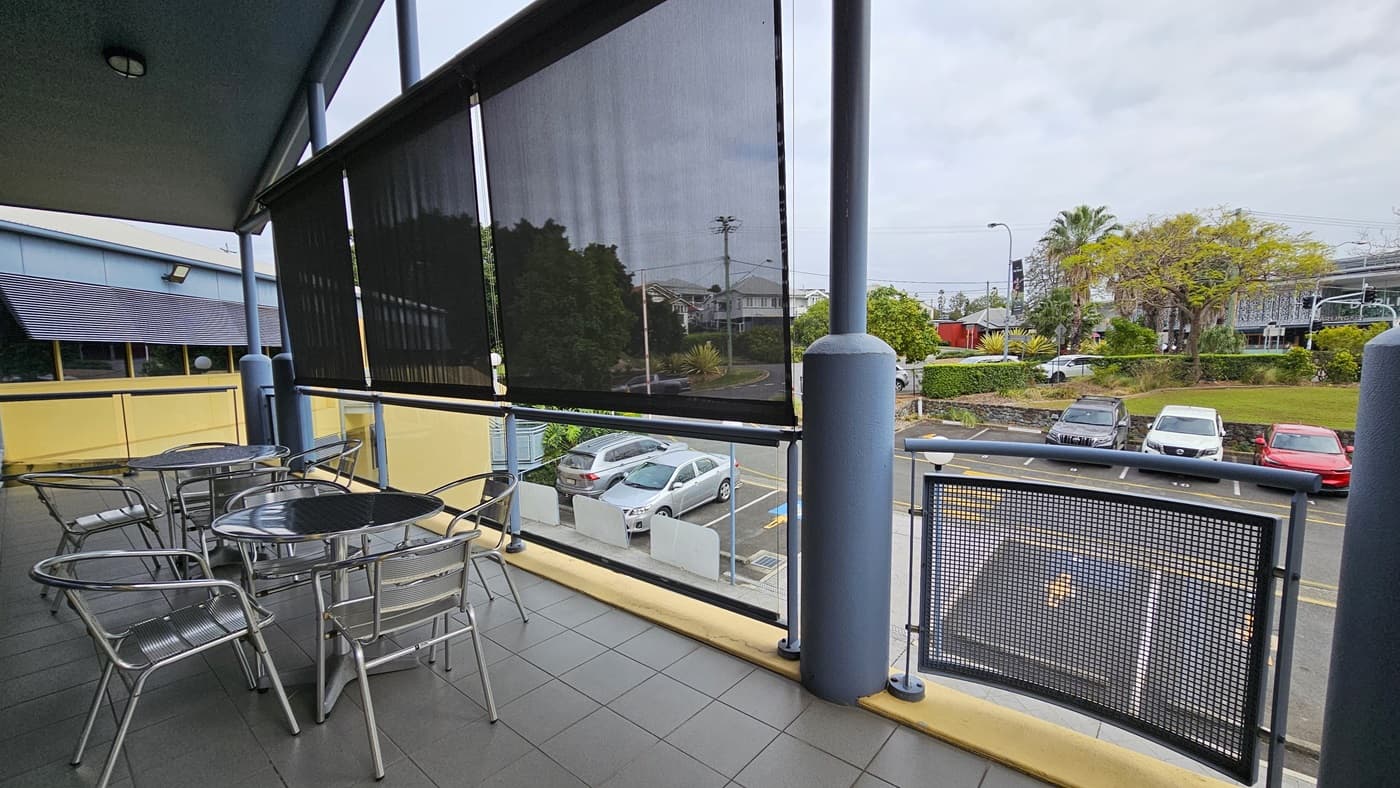 On the left, several round metal tables with matching chairs on a tiled balcony. In the middle, black roller blinds partially drawn, revealing a view of parked cars and a residential area in the background. On the right, a blue pillar and a metal railing with a mesh panel. The floor is covered with grey tiles. The ceiling is blue with a single light fixture.