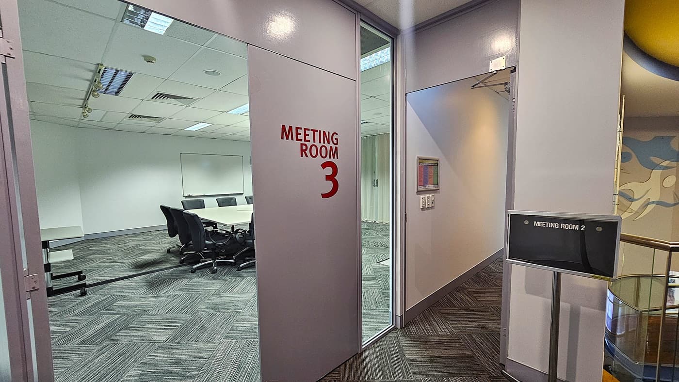 On the left, an open door labelled "Meeting Room 3" in red text, revealing a room with a long table and black office chairs. On the right, a sign for "Meeting Room 2" next to a hallway with grey patterned carpet tiles. In the background, a mural with abstract blue and white figures is partially visible. The floor is covered with grey patterned carpet tiles. The ceiling is white with recessed lighting, air vents, and acoustic tiles.