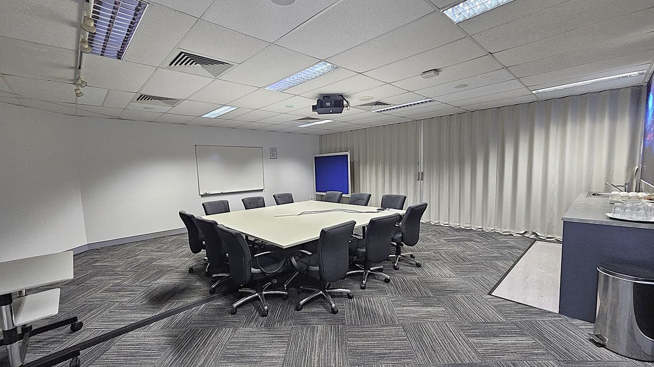 On the left, a whiteboard mounted on a white wall. In the middle, a large square table surrounded by black office chairs, with a ceiling-mounted projector above. On the right, a counter with a sink, cups, and plates, next to a large metal bin. The background features floor-to-ceiling white curtains. The floor is covered with grey patterned carpet tiles. The ceiling is white with recessed lighting, air vents, and acoustic tiles.