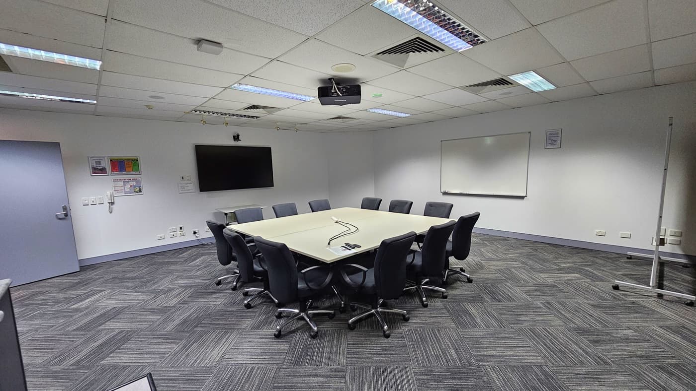 On the left, a closed grey door and a wall-mounted phone beneath a bulletin board. In the middle, a large square table surrounded by black office chairs, with a ceiling-mounted projector above. On the right, a whiteboard mounted on the wall. The floor is covered with grey patterned carpet tiles. The ceiling is white with recessed lighting, air vents, and acoustic tiles.