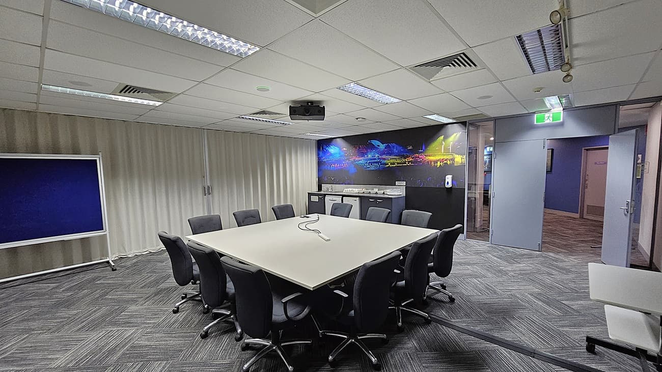 On the left, a blue noticeboard on a stand in front of floor-to-ceiling white curtains. In the middle, a large square table surrounded by black office chairs, with a ceiling-mounted projector above. On the right, a counter with a sink, cups, and plates, next to a mural of a stadium scene. The background includes an open door leading to a hallway. The floor is covered with grey patterned carpet tiles. The ceiling is white with recessed lighting, air vents, and acoustic tiles.
