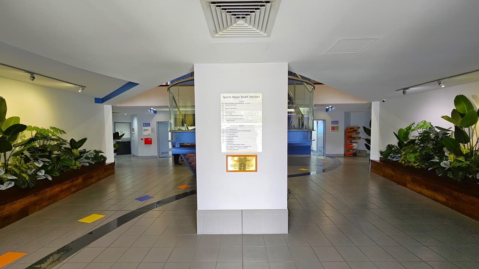 On the left, a yellow pillar with a security camera sign and a glass door with a notice. In the middle, a glass entrance with a mat on the tiled floor, leading to an interior with a central pillar displaying information. On the right, another yellow pillar with a mounted intercom and a glass door with a sign. The background includes a tiled walkway, a bench, and parked cars visible through the glass. The ceiling is white with recessed lighting.