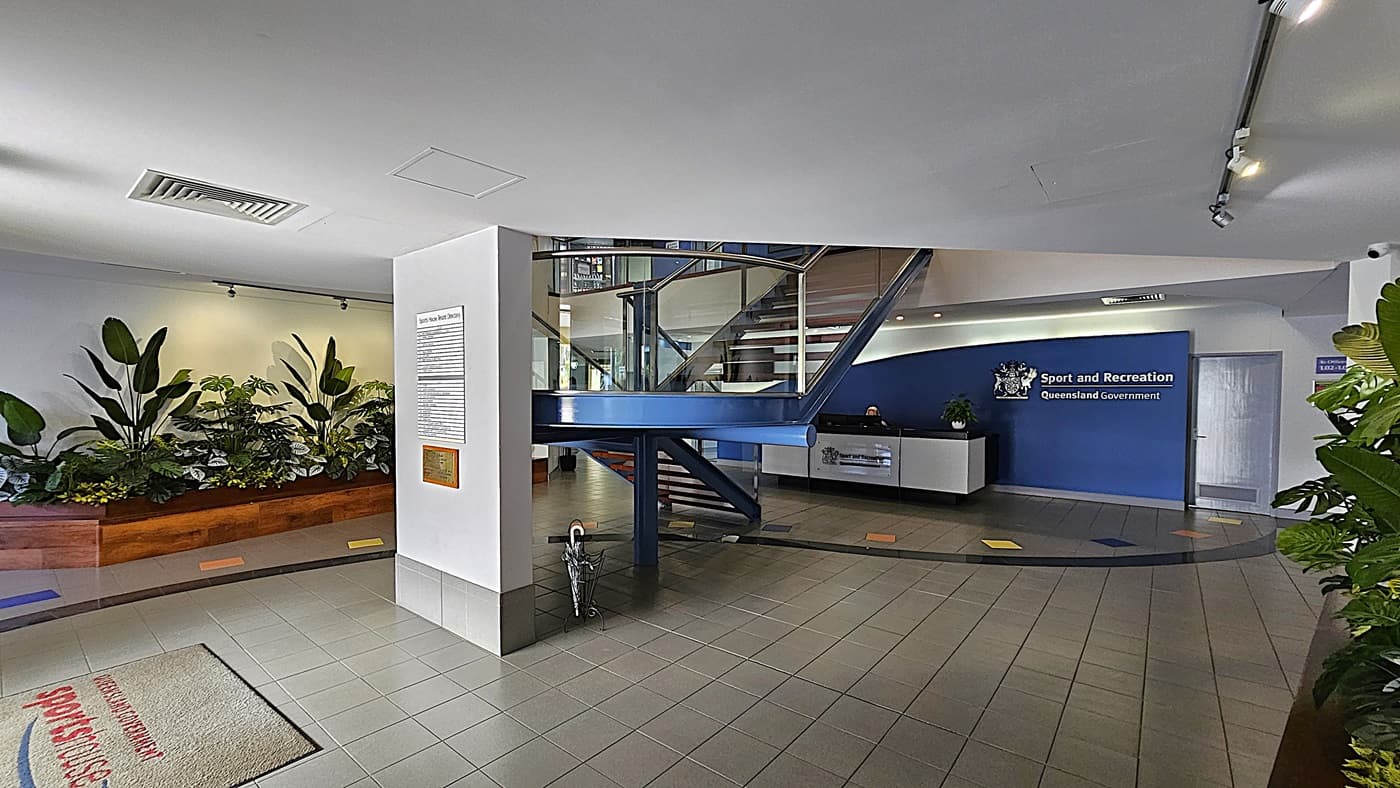 On the left, a wooden planter box with various green plants under track lighting. In the middle, a white pillar with a tenant directory sign and a gold plaque, flanked by a blue staircase with glass railings leading to the upper floor. On the right, a reception desk with a sign reading "Sport and Recreation Queensland Government" on a blue wall. The background includes a door and more plants. The floor is tiled with grey tiles and colourful square accents.