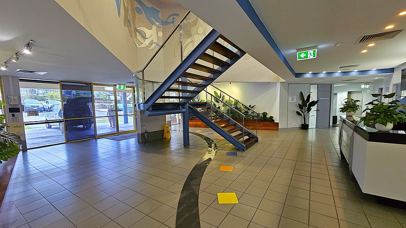 On the left, large glass doors with a view of parked cars outside and a yellow caution sign on the floor. In the middle, a blue staircase with wooden steps and glass railings leading to the upper floor, with a mural on the wall above. On the right, a reception desk with potted plants and a hallway with more plants and doors. The background includes a wooden planter box with green plants. The floor is tiled with grey tiles and colourful square accents. The ceiling is white with recessed lighting