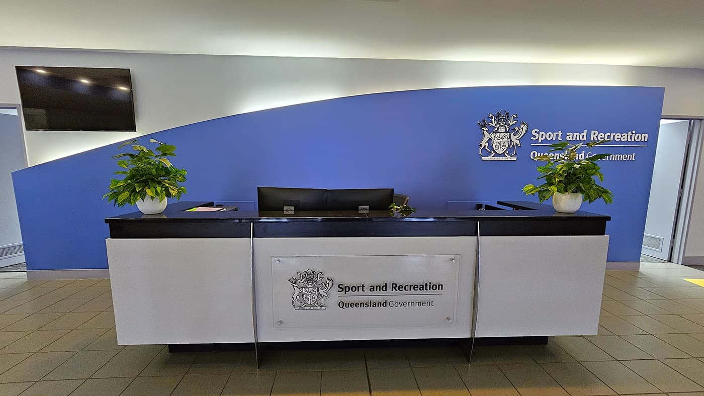 On the left, a wall-mounted flat-screen television above a doorway. In the middle, a black and white reception desk with two potted plants and a sign reading "Sport and Recreation Queensland Government" on the front. On the right, a blue wall with the same sign and a doorway leading to another room. The floor is tiled with beige tiles. The ceiling is white with recessed lighting and a curved design element above the desk.