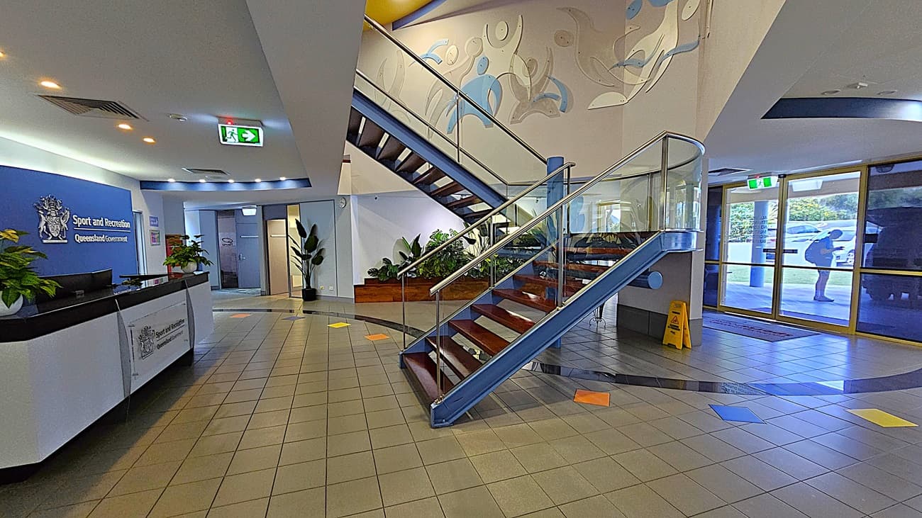 On the left, a reception desk with a sign reading "Sport and Recreation Queensland Government" on a blue wall, flanked by potted plants. In the middle, a staircase with wooden steps and glass railings, leading to an upper floor with a mural on the wall. On the right, large glass doors with a view of parked cars outside and a yellow caution sign on the floor. The floor is tiled with grey tiles and colourful square accents. The ceiling is white with recessed lighting and air vents.