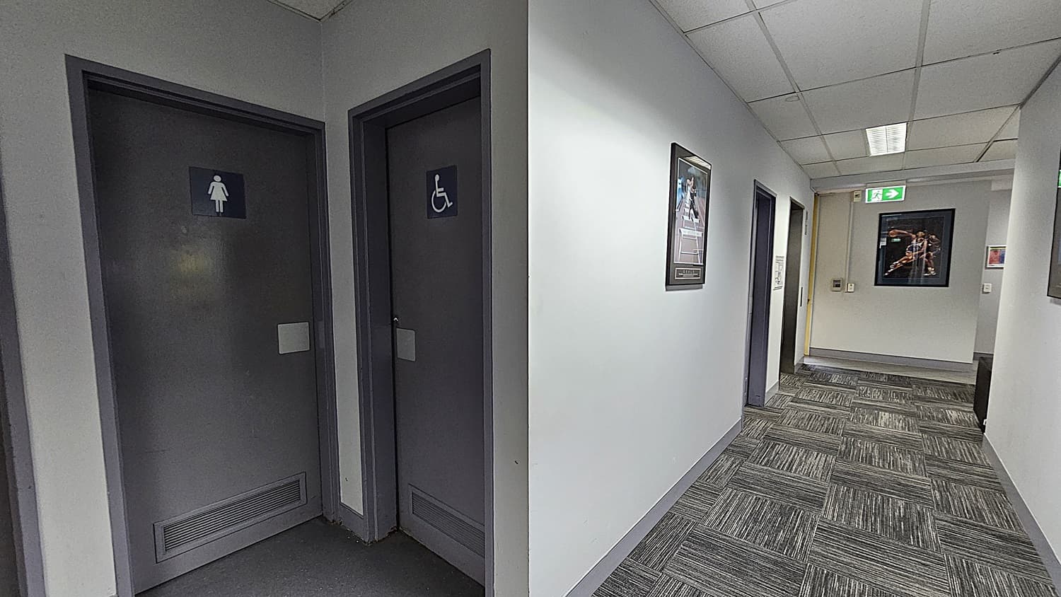 On the left, two grey doors with signs indicating a women's restroom and an accessible restroom. On the right, a white wall with framed pictures and a grey door. In the background, a hallway with more framed pictures, an emergency exit sign, and a doorway leading to another room. The floor is covered with grey patterned carpet tiles. The ceiling is white with a grid pattern and recessed fluorescent lighting.