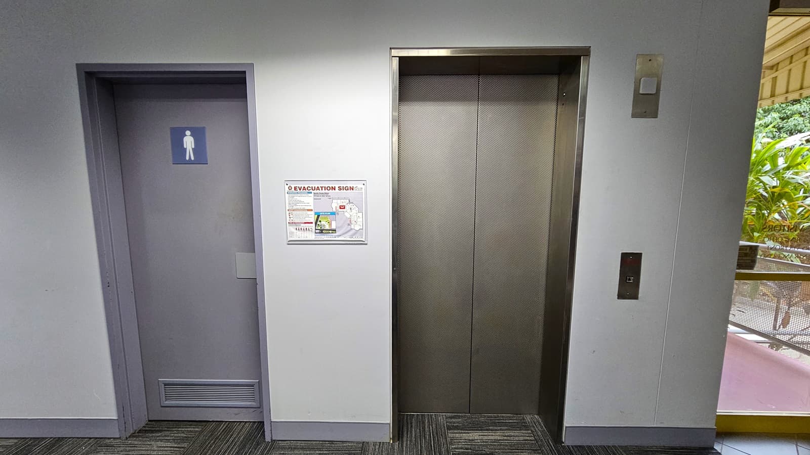 On the left, a grey door with a men's restroom sign. In the middle, a white wall with an evacuation sign and a stainless steel lift door. On the right, a window with a view of greenery outside and a yellow handrail. The floor is covered with grey patterned carpet tiles. The ceiling is white with recessed lighting.