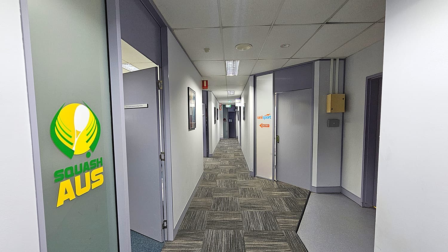 On the left, a frosted glass door with a "Squash AUS" logo in green and yellow, next to an open grey door. On the right, a grey door with a "UniSport" and "QUT Sport" logo, and a yellow electrical box mounted on the wall. In the background, a long hallway with framed pictures on the walls, a fire extinguisher, and an emergency exit sign. The floor is covered with grey patterned carpet tiles. The ceiling is white with a grid pattern and recessed fluorescent lighting.