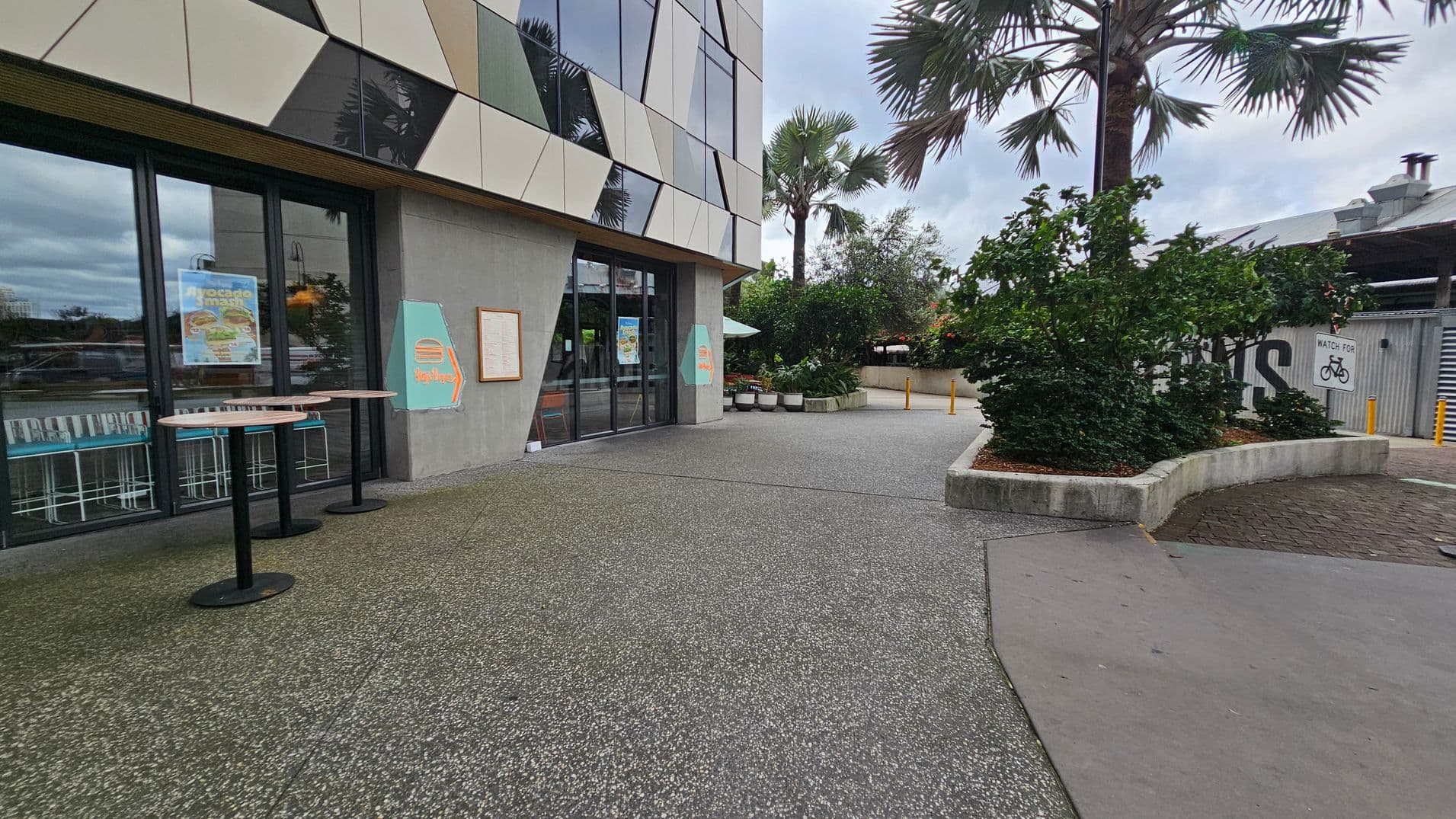 A modern building with geometric patterns on the facade is on the left, featuring large glass doors and posters. Two round tables stand outside. The ground is a mix of green and grey textured surfaces. On the right, there are landscaped areas with bushes and a palm tree. A sign warns to watch for bicycles. In the background, there is a metal structure with a partially visible word. The sky is overcast.