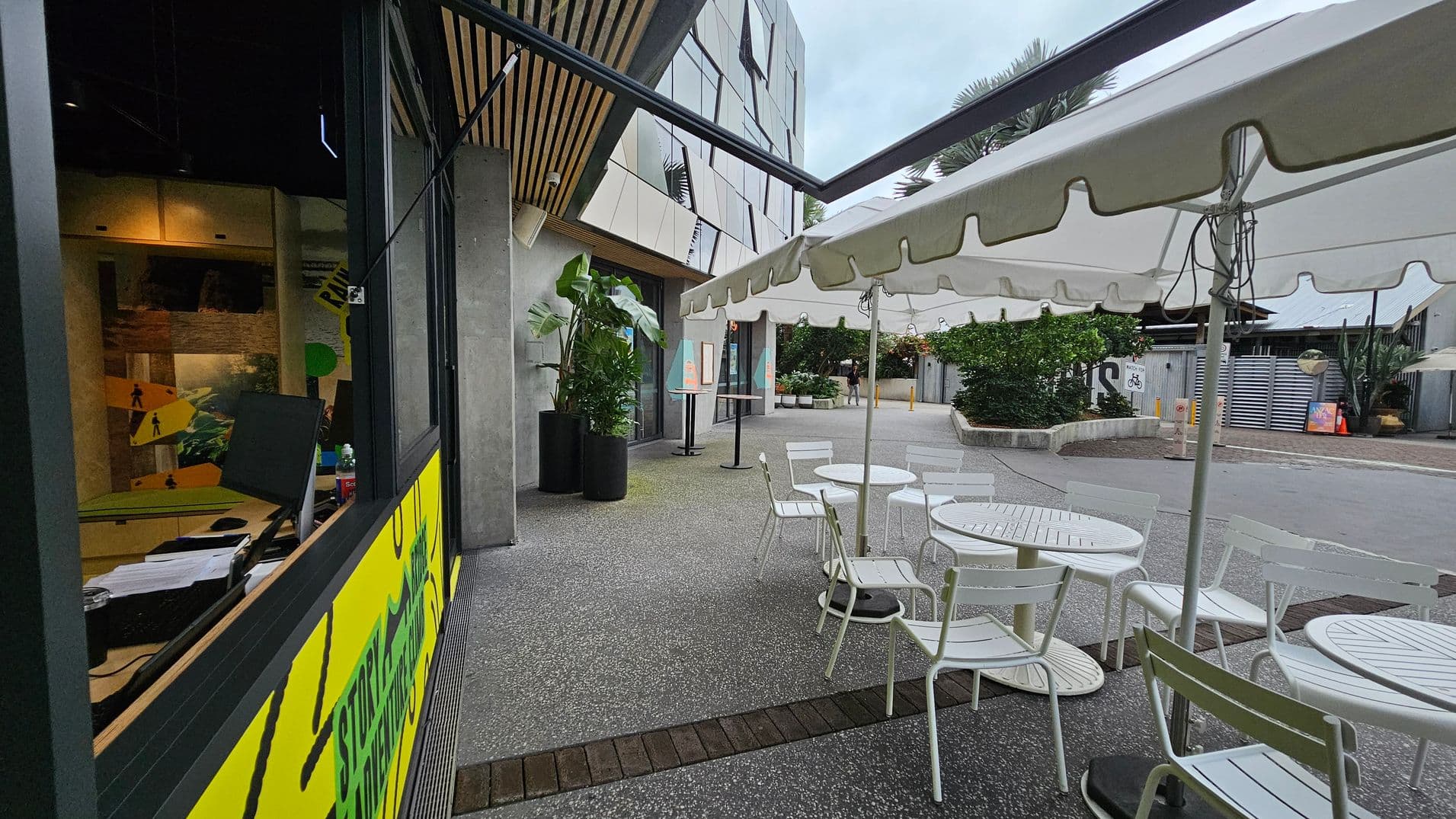 On the left, there's a window with a yellow and green sign, and inside, a desk with a computer monitor and papers. The ceiling has wooden slats. In the middle, there are white tables and chairs under a white canopy. On the right, a pathway leads to a building with a modern facade. Potted plants are near the entrance, and a few people are walking in the distance. The ground is paved with a mix of concrete and brick.