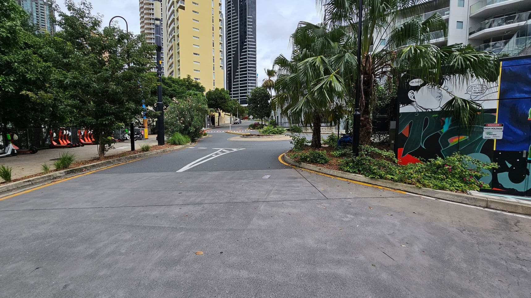 A paved pathway with a slight curve, flanked by greenery and palm trees. On the left, there are parked electric scooters and a row of trees. On the right, a mural with a comic-style illustration is visible on a wall, accompanied by a "Danger High Voltage" sign. In the background, tall buildings rise, including a yellow one and a dark, modern skyscraper. The area is bordered by low shrubs and plants.
