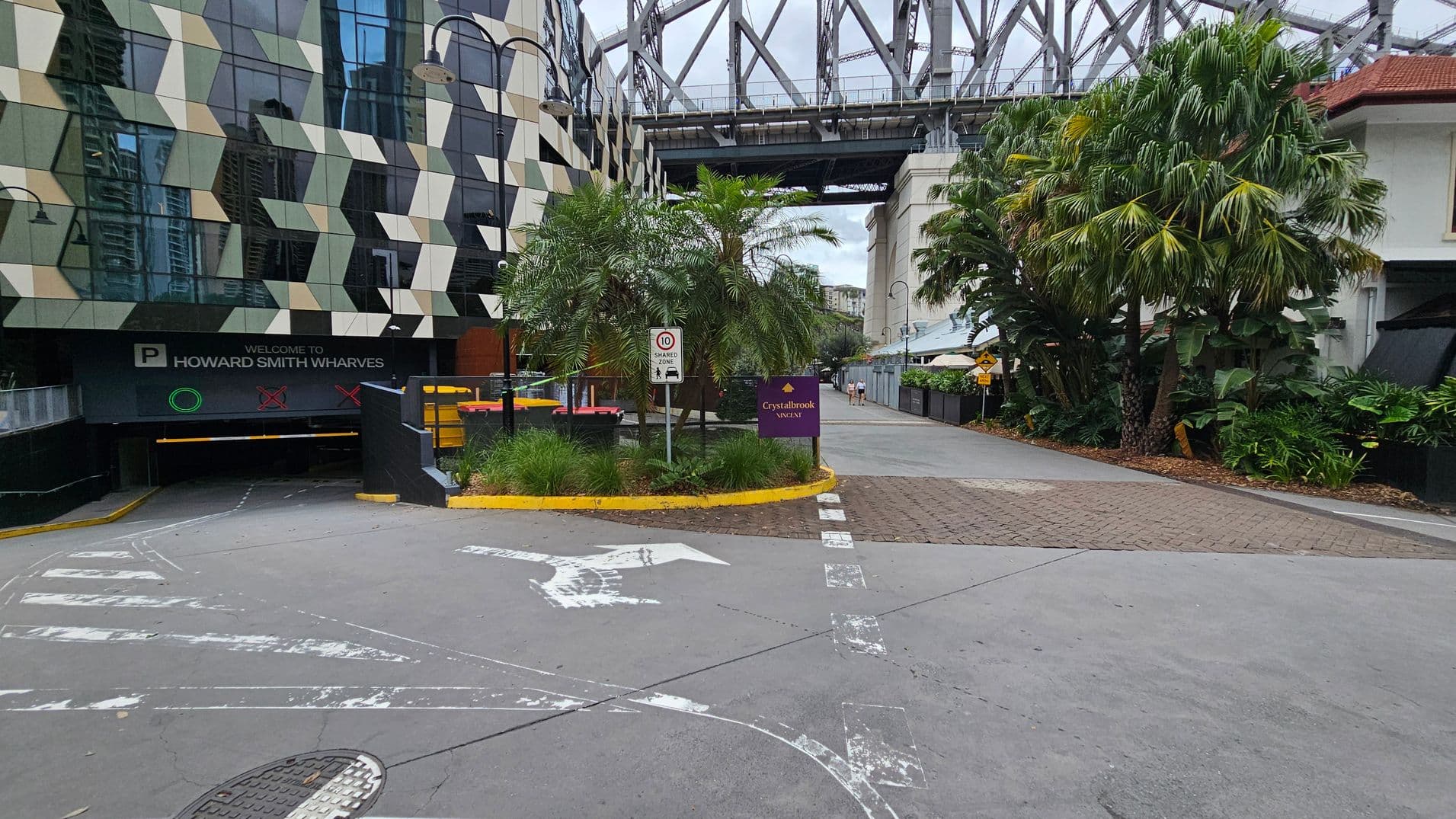 On the left, a modern building with a geometric pattern of beige, grey, and black panels. Below it, a sign reads "Welcome to Howard Smith Wharves" above a parking entrance. In the centre, a small landscaped area with palm trees and a sign for "Crystalbrook Vincent." On the right, a pathway lined with lush greenery and more palm trees. In the background, a large steel bridge structure spans across the scene. The ground is a mix of concrete and brick paving.