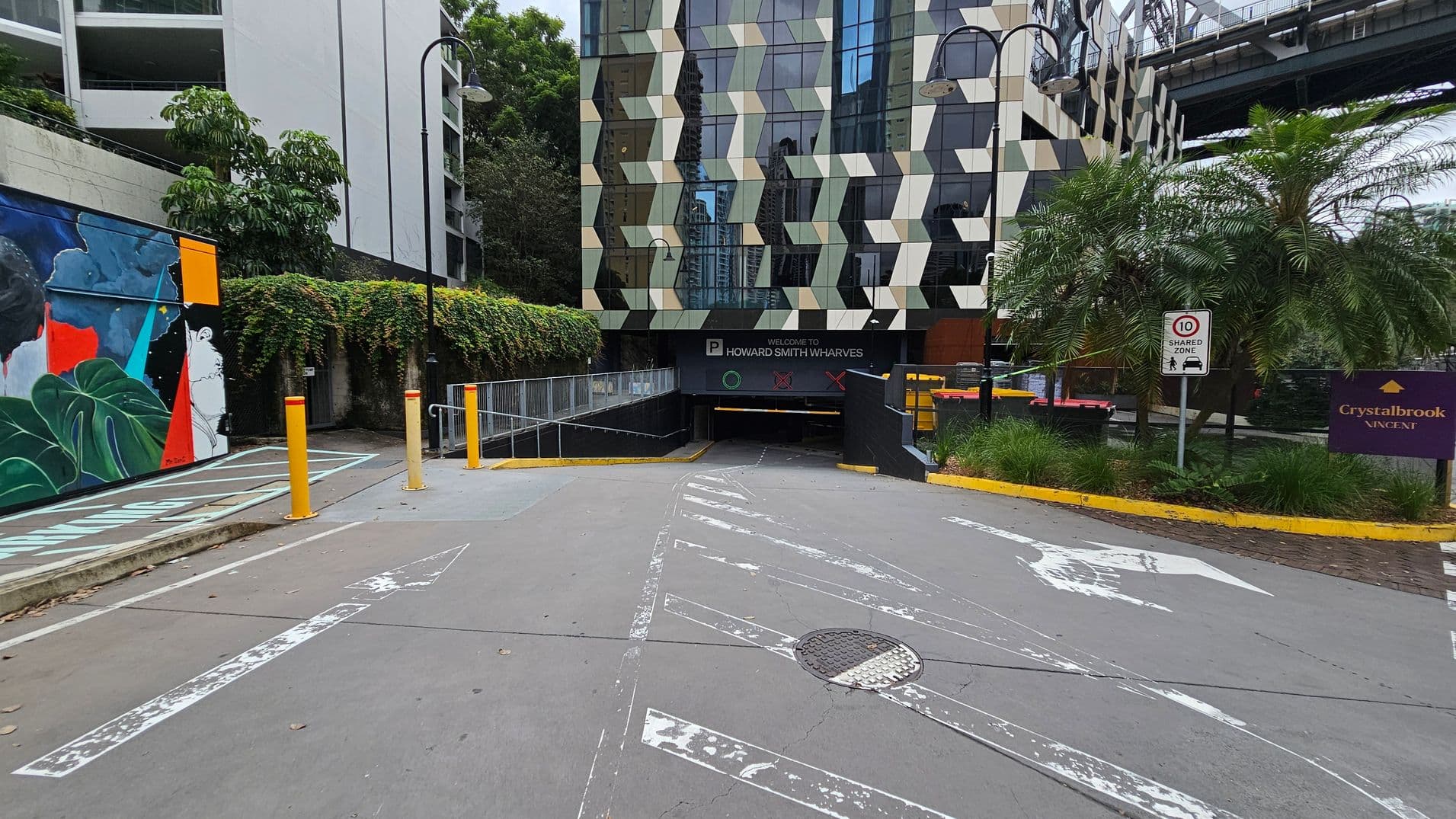 Entrance to an underground car park with a geometric-patterned building in the background. The left side features a colourful mural and greenery, while the right side has palm trees and a sign indicating a shared zone with a speed limit of 10. The ground is marked with directional arrows, and there are yellow bollards near the entrance. A sign reads "Welcome to Howard Smith Wharves" above the car park entrance.