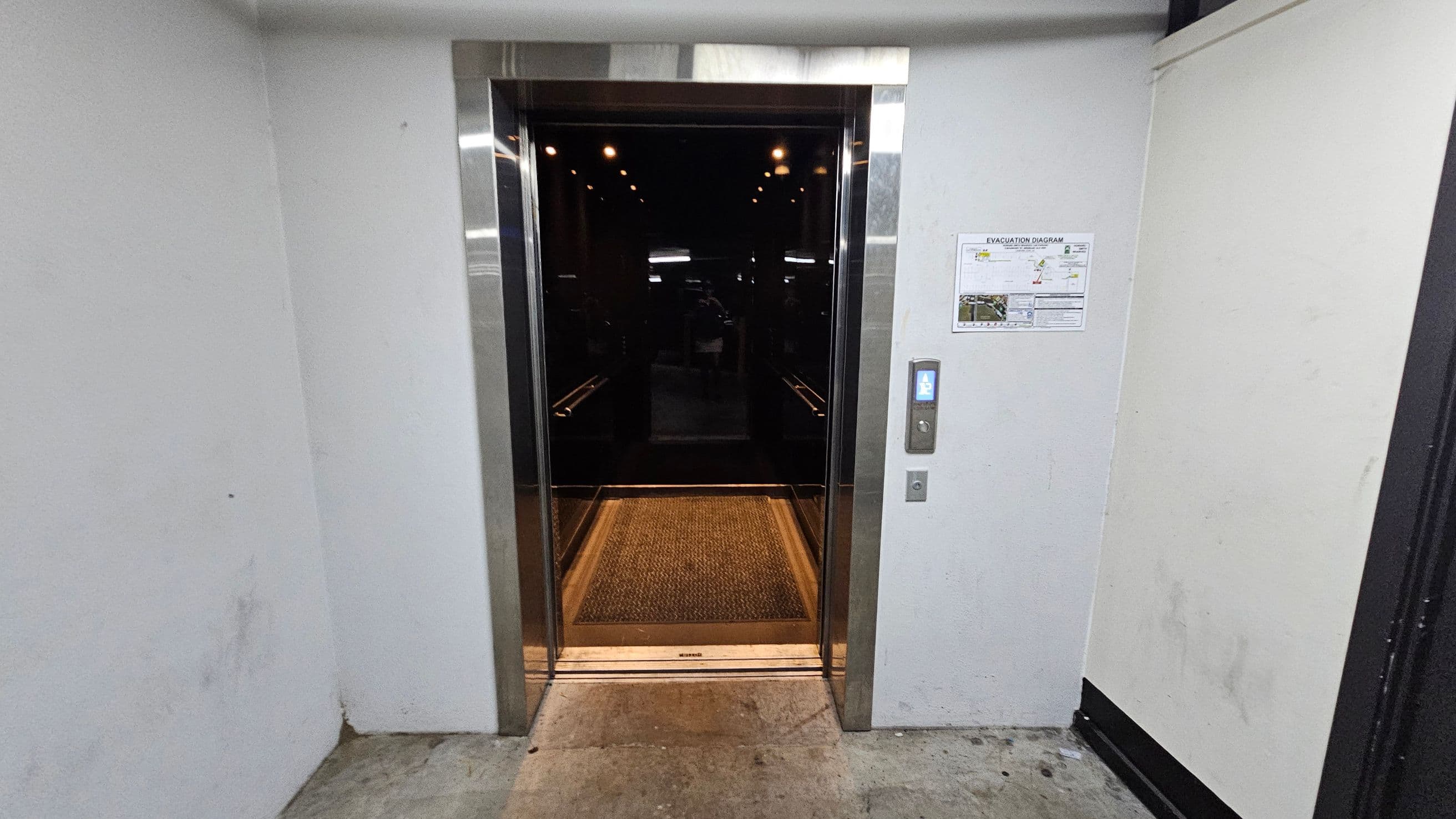 An open lift with shiny metal doors and a dark interior. The floor inside the lift is covered with a textured mat. On the right wall outside the lift, there is a control panel with buttons and a lit display, along with an evacuation diagram posted above it. The surrounding walls are plain and light-coloured, with a slightly worn floor in front of the lift entrance.