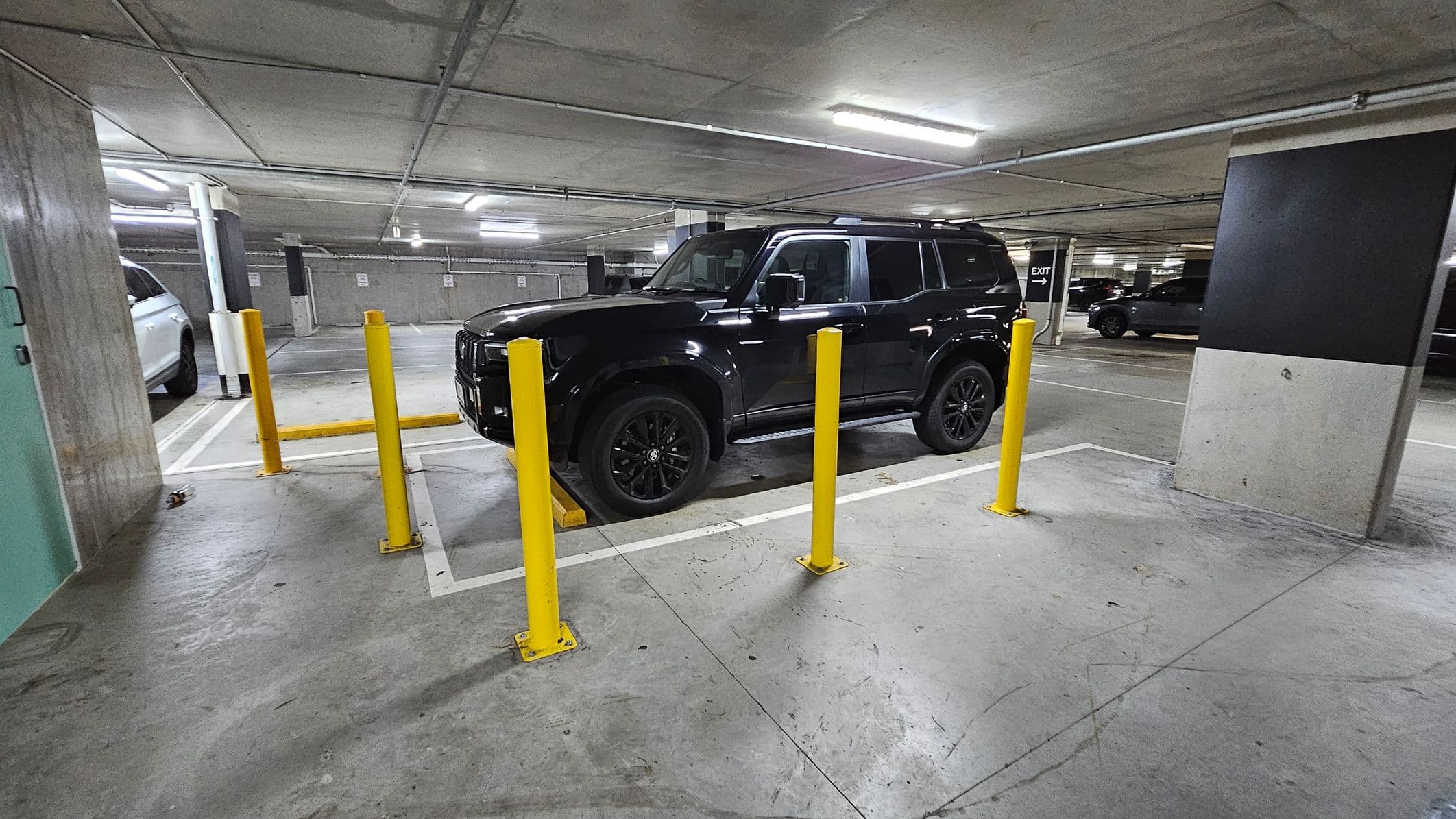A black SUV is parked in a basement car park, surrounded by yellow bollards. The floor is concrete, and the ceiling is low with fluorescent lighting. To the left, a white vehicle is partially visible. In the background, there are more parked cars and a sign indicating the exit. The area is enclosed by concrete walls and pillars.