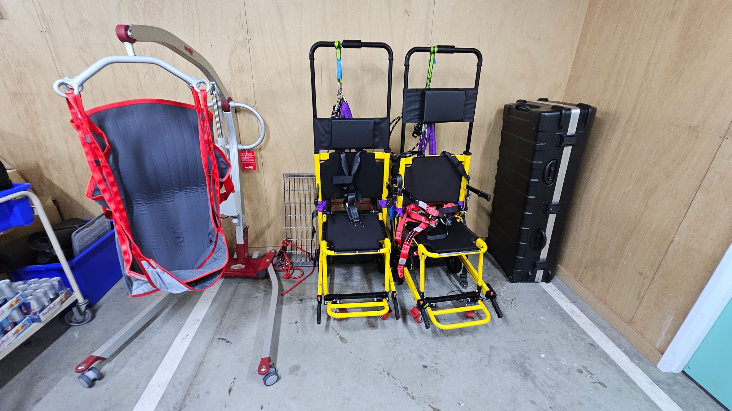 On the left, there's a red and grey patient lift with a sling, next to a trolley with blue bins and a pack of canned drinks. In the centre, two yellow evacuation chairs with black seats and red and purple straps are positioned side by side. On the right, a large black case is standing upright against a wooden wall. The floor is concrete with visible markings.