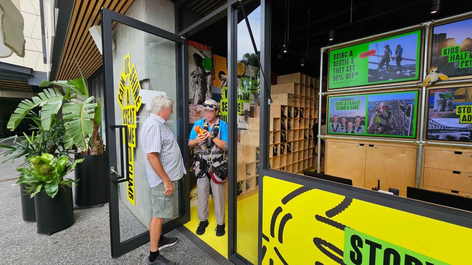 A man in a checkered shirt stands at the entrance of a shop, talking to another man in climbing gear. The shop has a glass door with a yellow sign that reads "Rain or Shine, A Great Climb." Inside, there are wooden cubbies and promotional posters offering discounts for climbing activities. Large potted plants are placed outside on the left. The floor inside is yellow, and the ceiling has wooden slats.