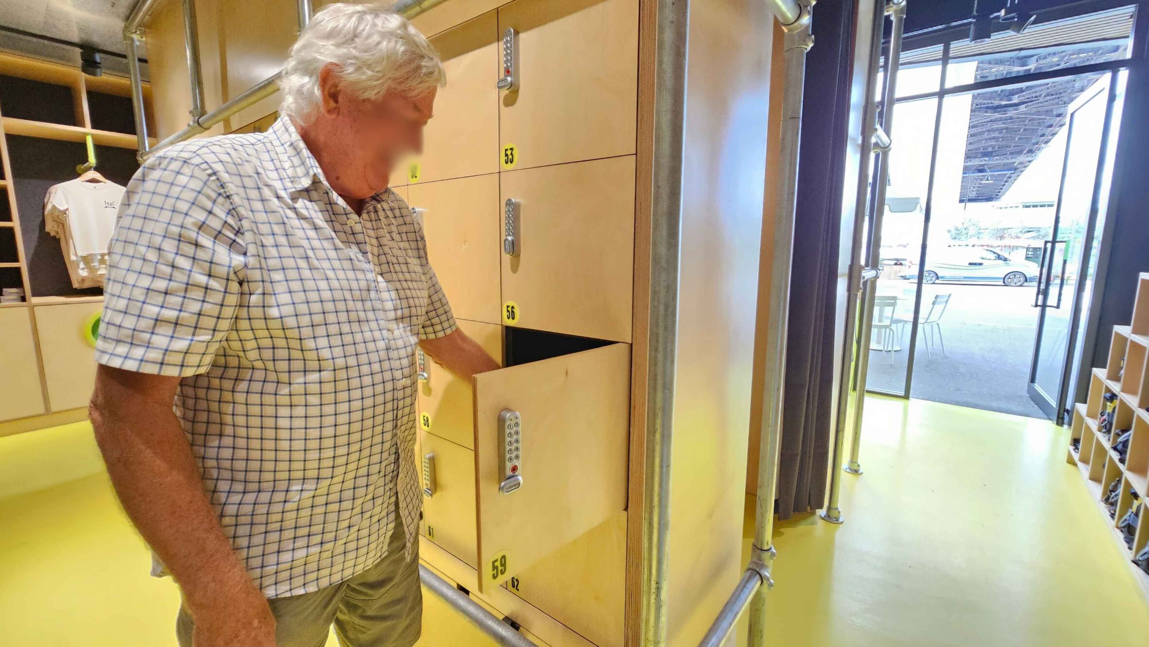 A man with grey hair wearing a checked shirt is standing on a yellow floor, opening a locker with the number 56. The lockers are light-coloured with combination locks. To the right, there is a glass door leading outside, where a car and some outdoor seating are visible. The ceiling has exposed pipes, and there are shelves with items on the left.