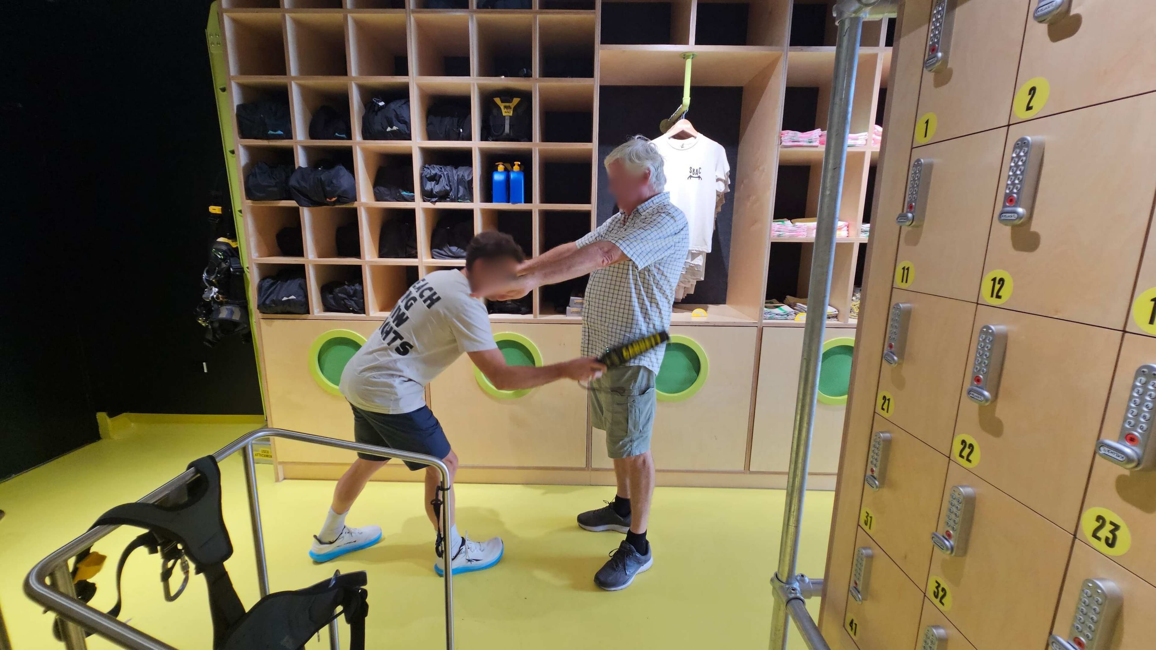 A man in a white T-shirt and shorts is bending forward, holding a device near another man in a checked shirt and shorts. They are in front of a wooden shelving unit filled with black bags, water bottles, and a hanging T-shirt. To the right, there are numbered lockers with combination locks. The floor is bright yellow, and there is a metal railing in the foreground.