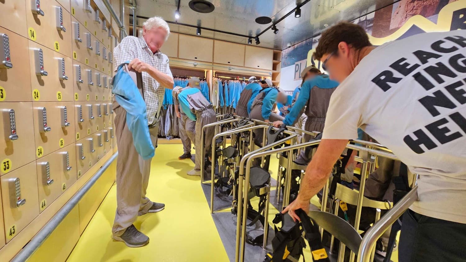 A room with a bright yellow floor and a ceiling with spotlights. On the left, there are beige lockers with numbered yellow labels. In the centre, a man in a checked shirt is holding a blue garment. On the right, another man in a white T-shirt is handling climbing harnesses. In the background, several people are wearing blue and grey uniforms, preparing equipment. The walls have racks of similar blue garments.