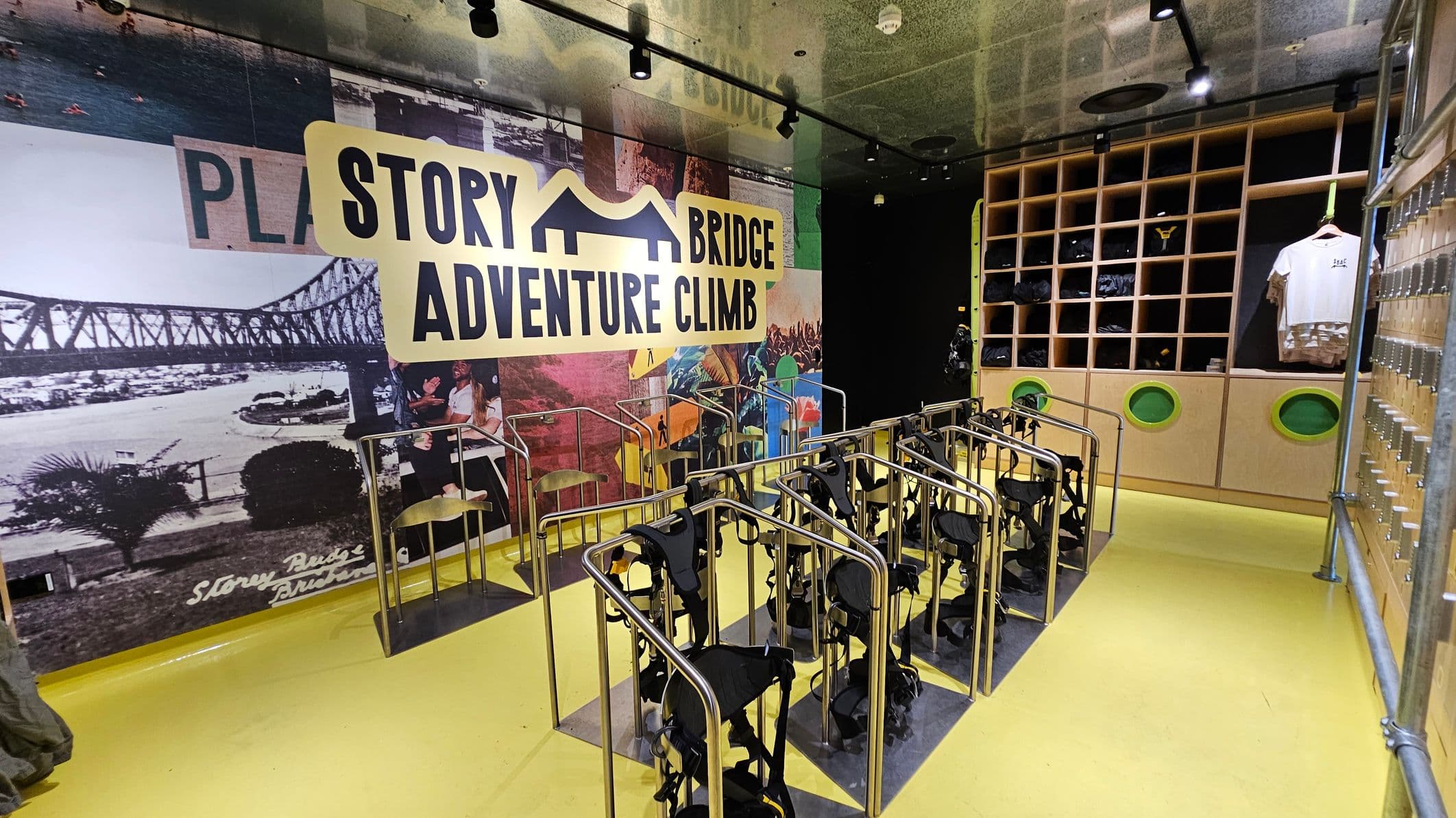 A room with a bright yellow floor and a ceiling with spotlights. On the left, a large mural features the words "Story Bridge Adventure Climb" and an image of a bridge. In the centre, several harnesses are neatly arranged on metal racks. On the right, a wooden shelving unit holds various items, including a white T-shirt on display. The wall also has small lockers with combination locks.
