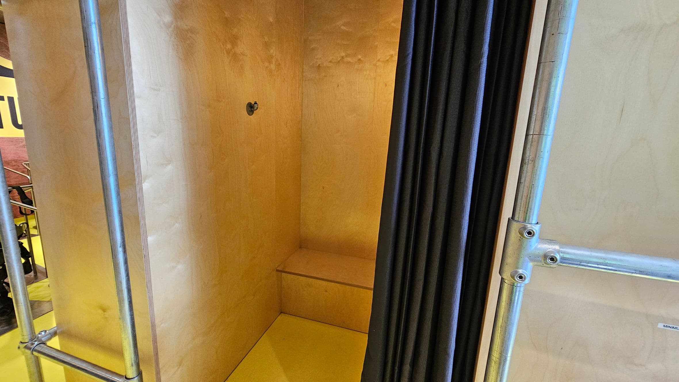 A small wooden booth with a light-coloured interior and a simple bench. The booth is framed by metal pipes on the left and right. A black curtain partially covers the entrance on the right side. The floor inside the booth is the same light wood as the walls. In the background, there is a hint of a yellow wall with some text partially visible.
