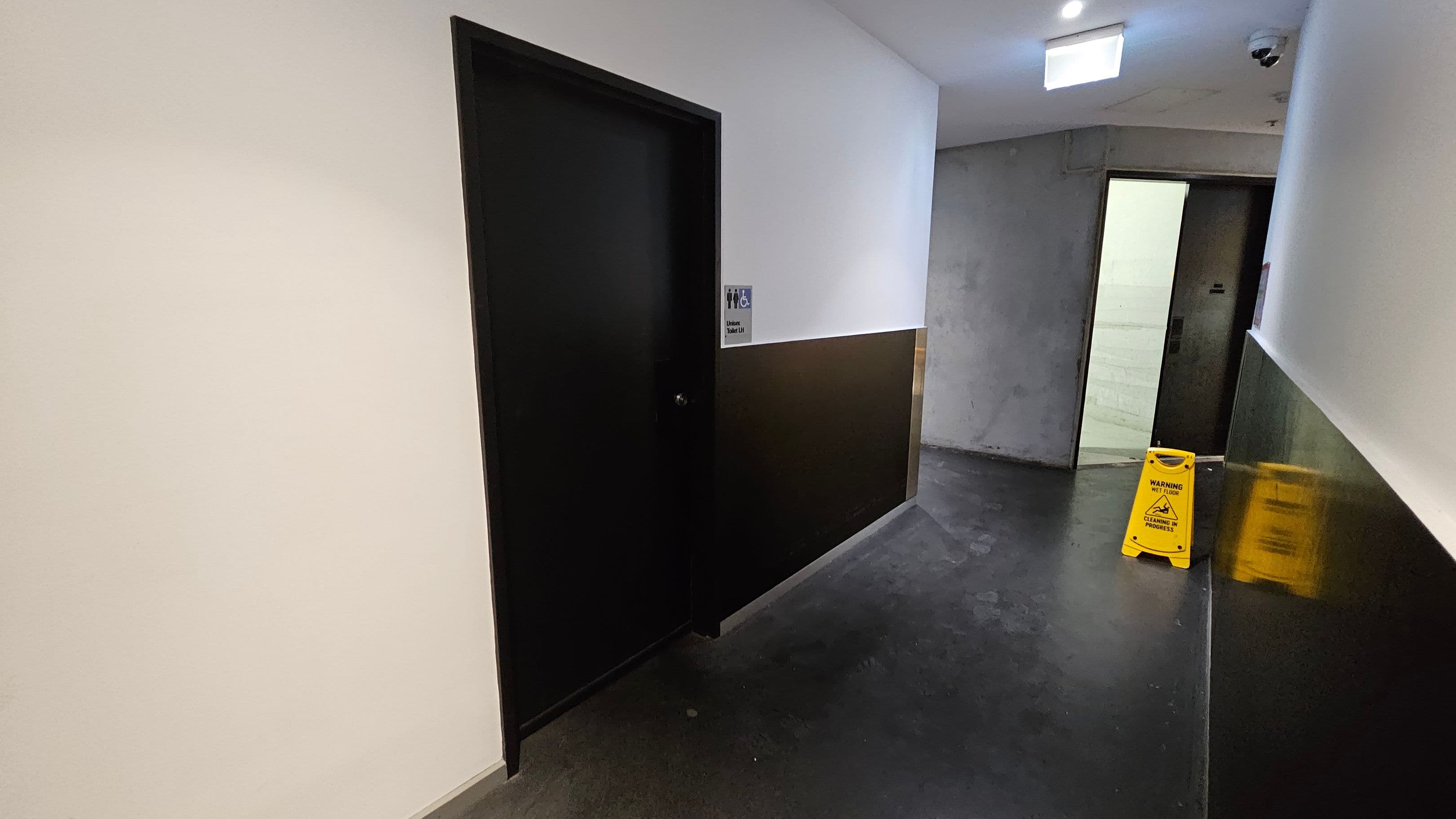 A corridor with a dark floor and white ceiling. On the left, a black door with a sign indicating a unisex and accessible toilet. On the right, a grey wall with an open doorway leading to a lit room. A yellow caution sign is placed on the floor near the doorway. A security camera is mounted on the ceiling above the doorway.