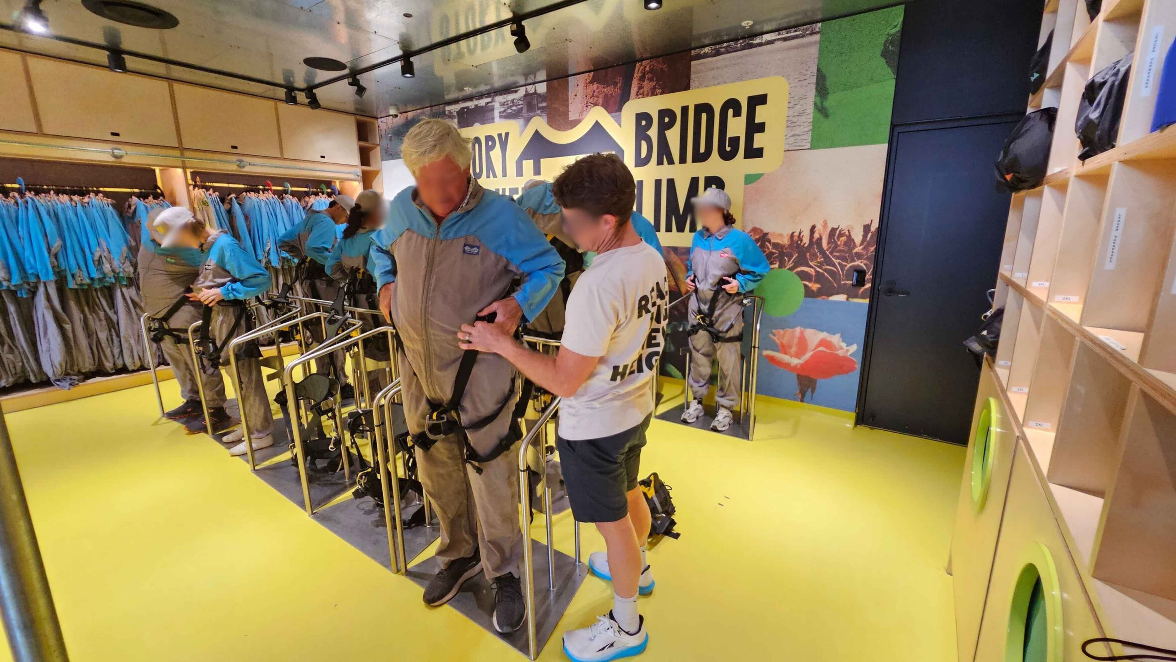 In a room with a bright yellow floor and a metallic ceiling, several people are preparing for an activity. On the left, individuals are wearing blue and grey suits, adjusting harnesses. In the centre, a person is being assisted with their harness by another individual in a white shirt. The background features a colourful mural with the words "BRIDGE CLIMB" and images of nature. On the right, there are shelves with bags and a closed door.