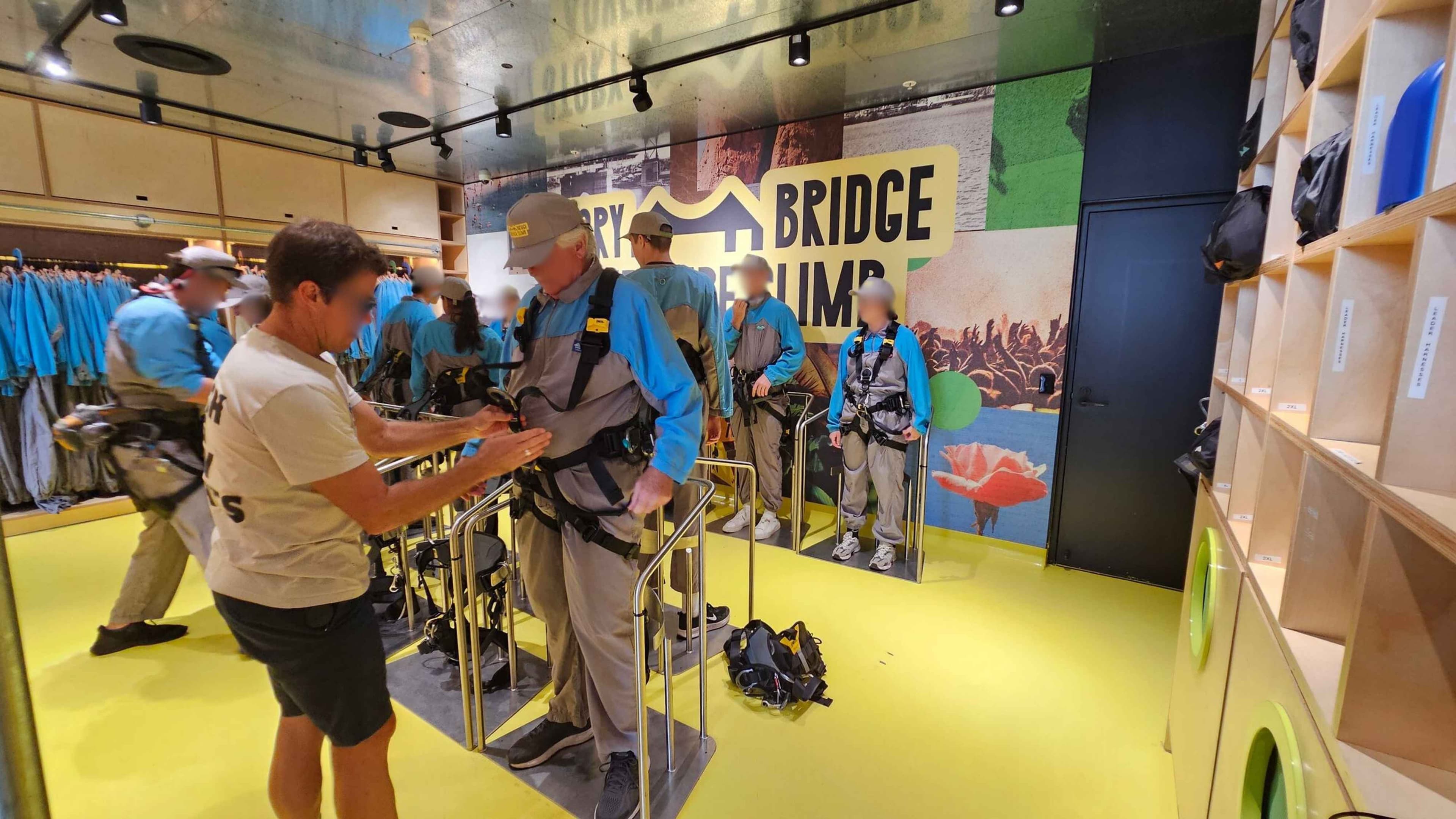 In a room with a bright yellow floor and a metallic ceiling, several people are preparing for an activity. On the left, a person in a beige shirt assists another in a blue and grey outfit with a harness. In the background, more individuals in similar attire stand near a wall with a large "Bridge Climb" sign. On the right, there are wooden cubbies with bags. The atmosphere suggests a preparation area for an outdoor adventure.