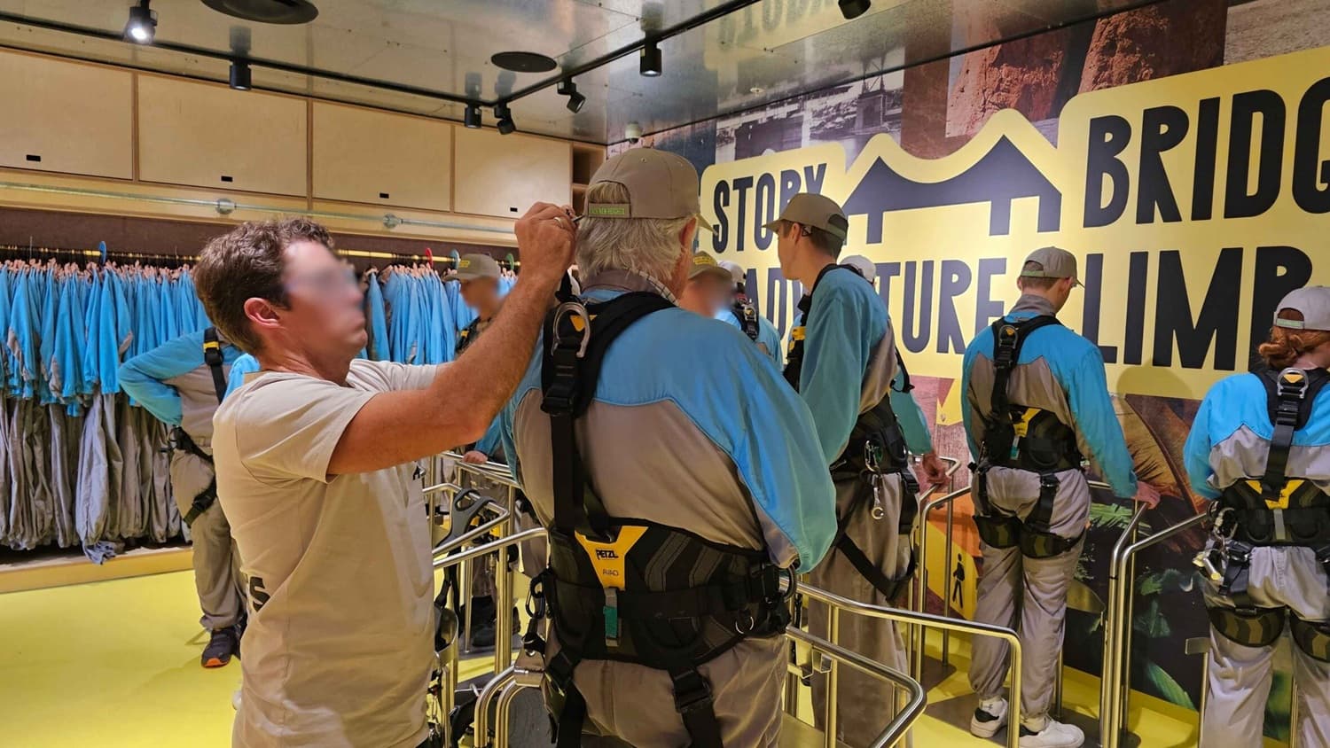 In a room with a yellow floor and a ceiling with spotlights, several people are wearing blue and grey climbing suits with harnesses. On the left, a man in a beige shirt adjusts the harness of another person. Behind them, racks of blue and grey suits hang. On the right, a large yellow and black sign reads "BRIDGE CLIMB" with a bridge icon. The group stands near metal railings, preparing for an activity.