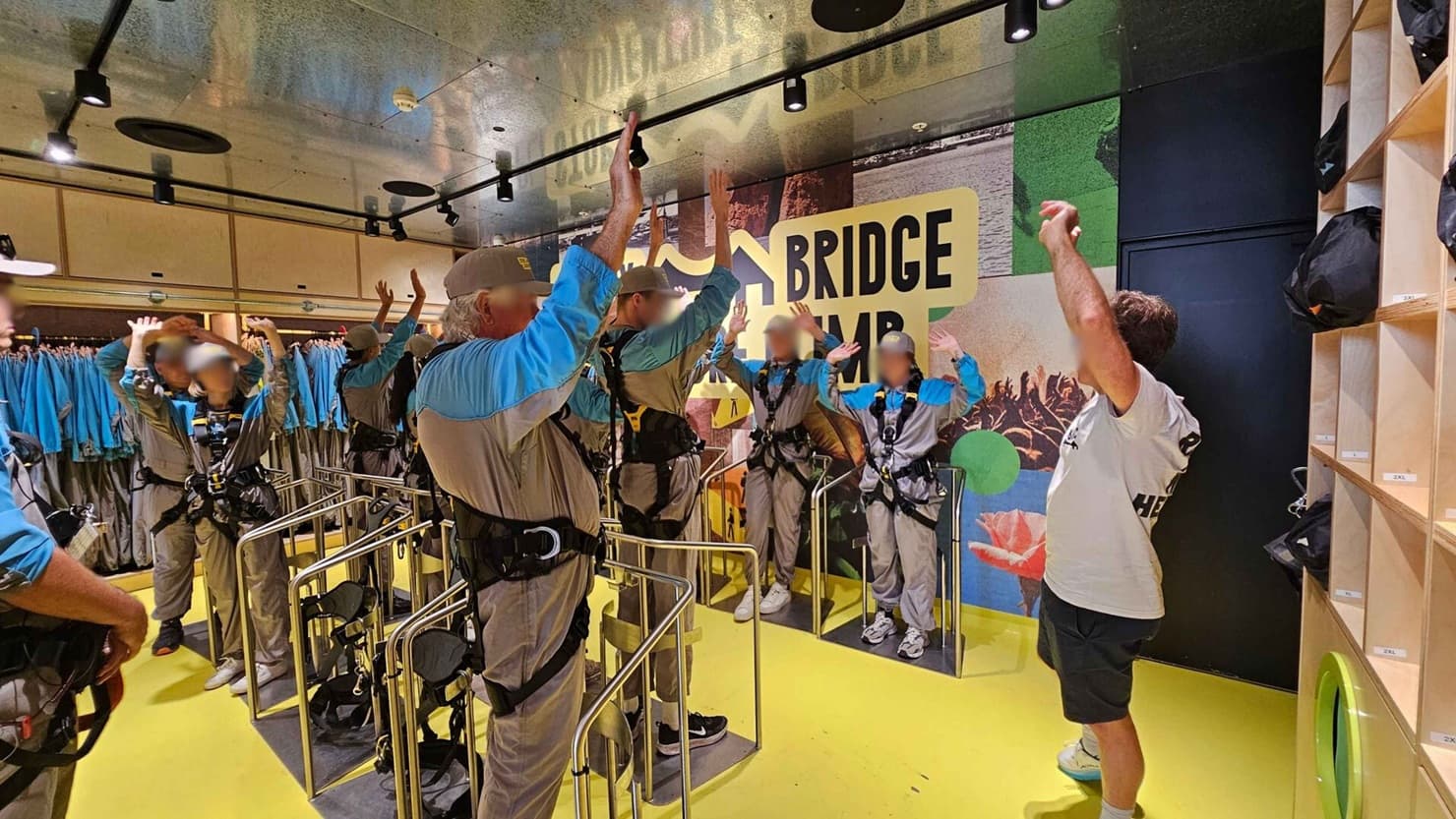 A group of people wearing harnesses and helmets stand in a room with a bright yellow floor. They are raising their arms, possibly in preparation for an activity. On the right, a person in a white shirt appears to be instructing them. The background features a large sign with the words "BRIDGE CLIMB" and colourful wall graphics. On the left, there are racks of blue and grey suits, and on the right, shelves with black bags. The ceiling is reflective with spotlights.